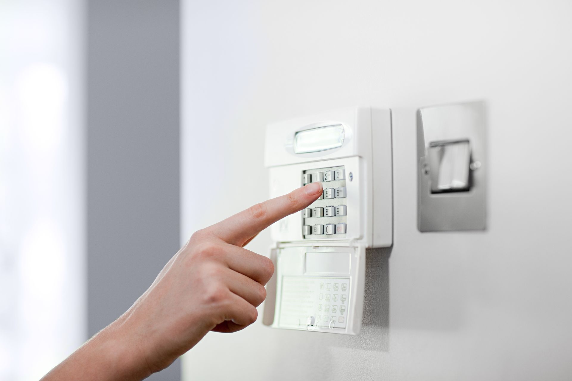 Person's finger pressing numbers on a white security system keypad mounted on a white wall.