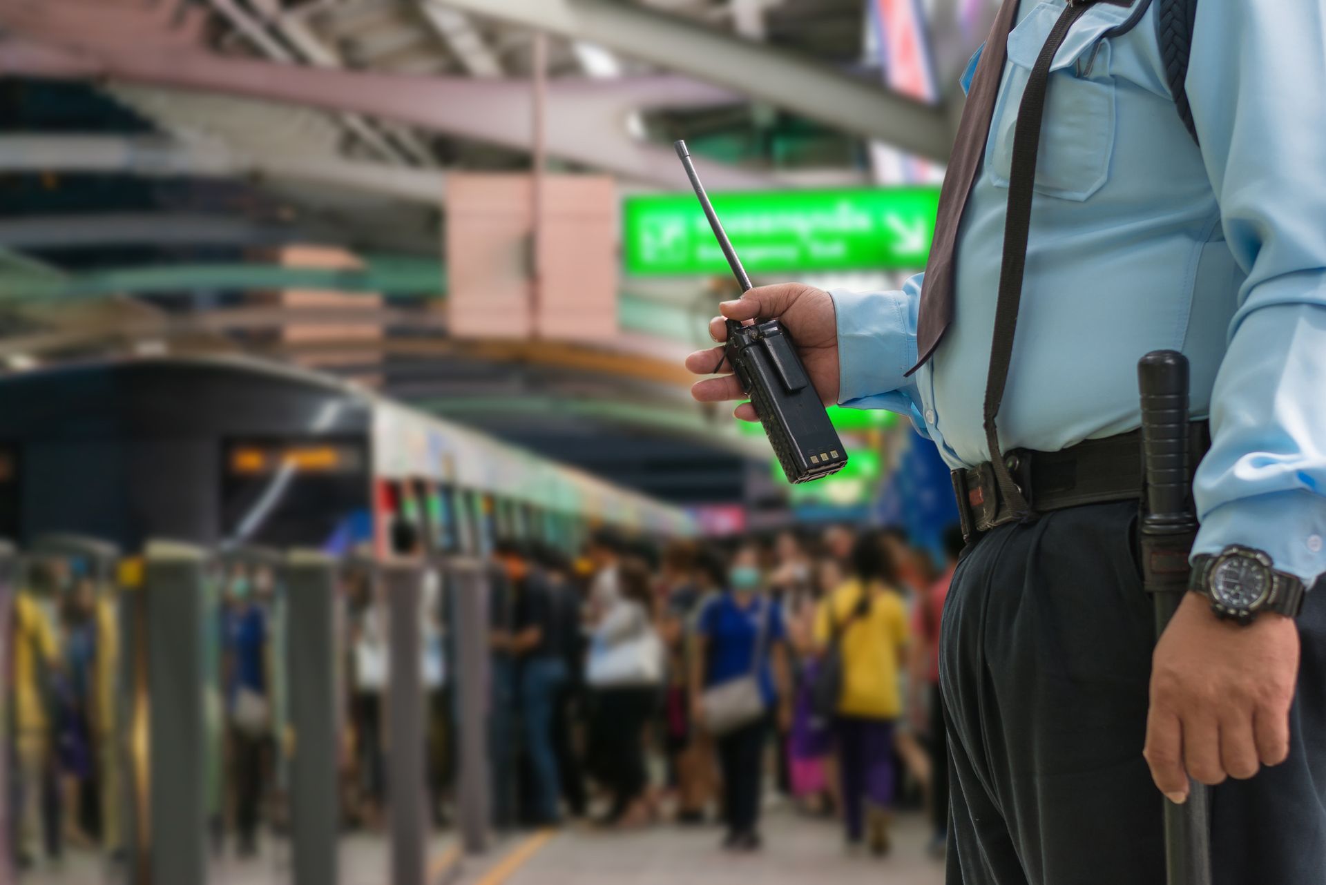 Security guard holding a walkie-talkie and baton stands at a train station entrance, watching the crowd.