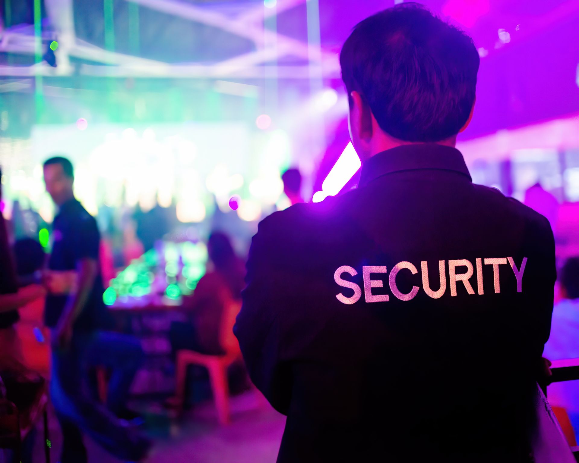 Security guard in black uniform watches a crowded, lit nightclub.