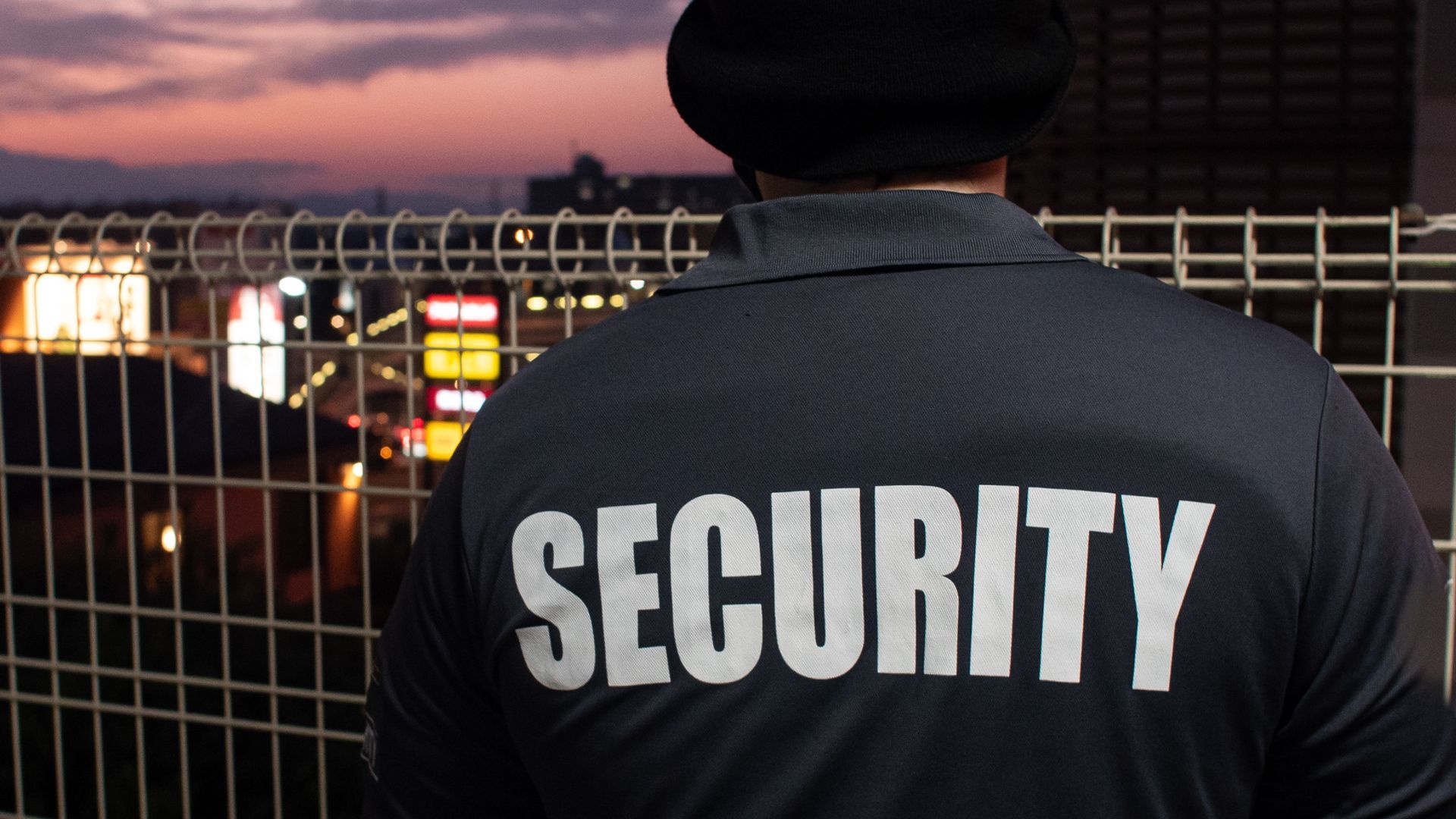 Security guard, back turned, overlooking a city at dusk. 