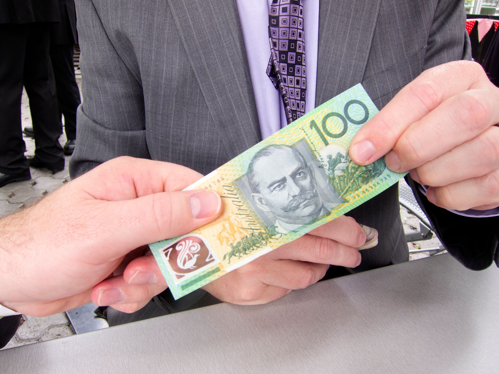 Person in a suit holding an Australian $100 note, with another person's hands visible.