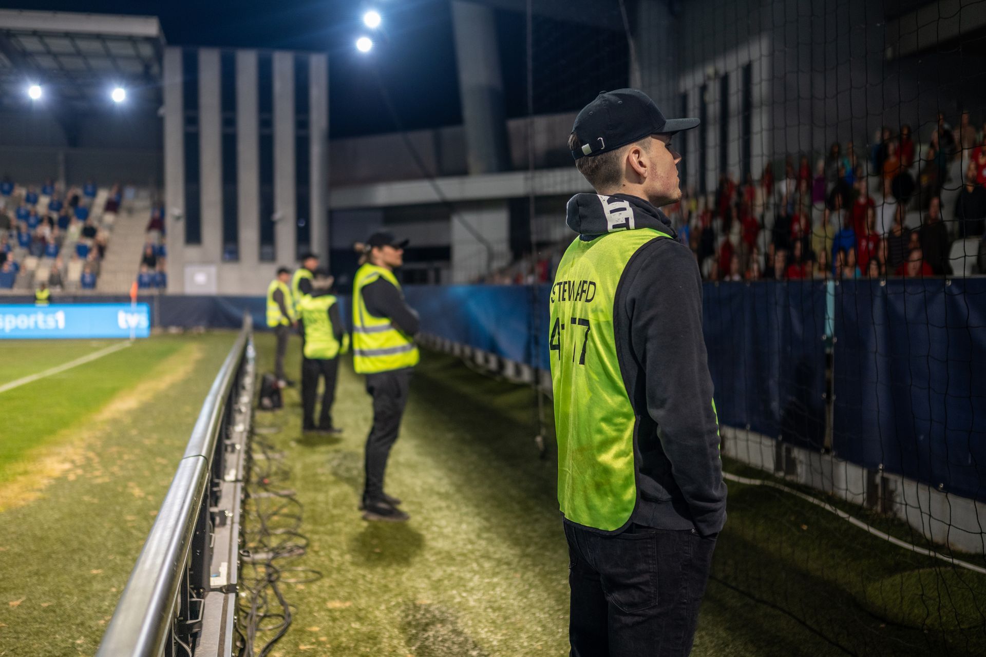 Security personnel in neon vests at a sports stadium, overseeing a game at night.
