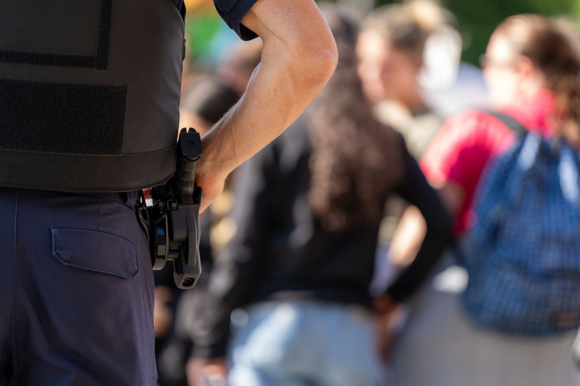 Police officer with a holstered gun, wearing a vest, standing in front of a crowd of blurred people.