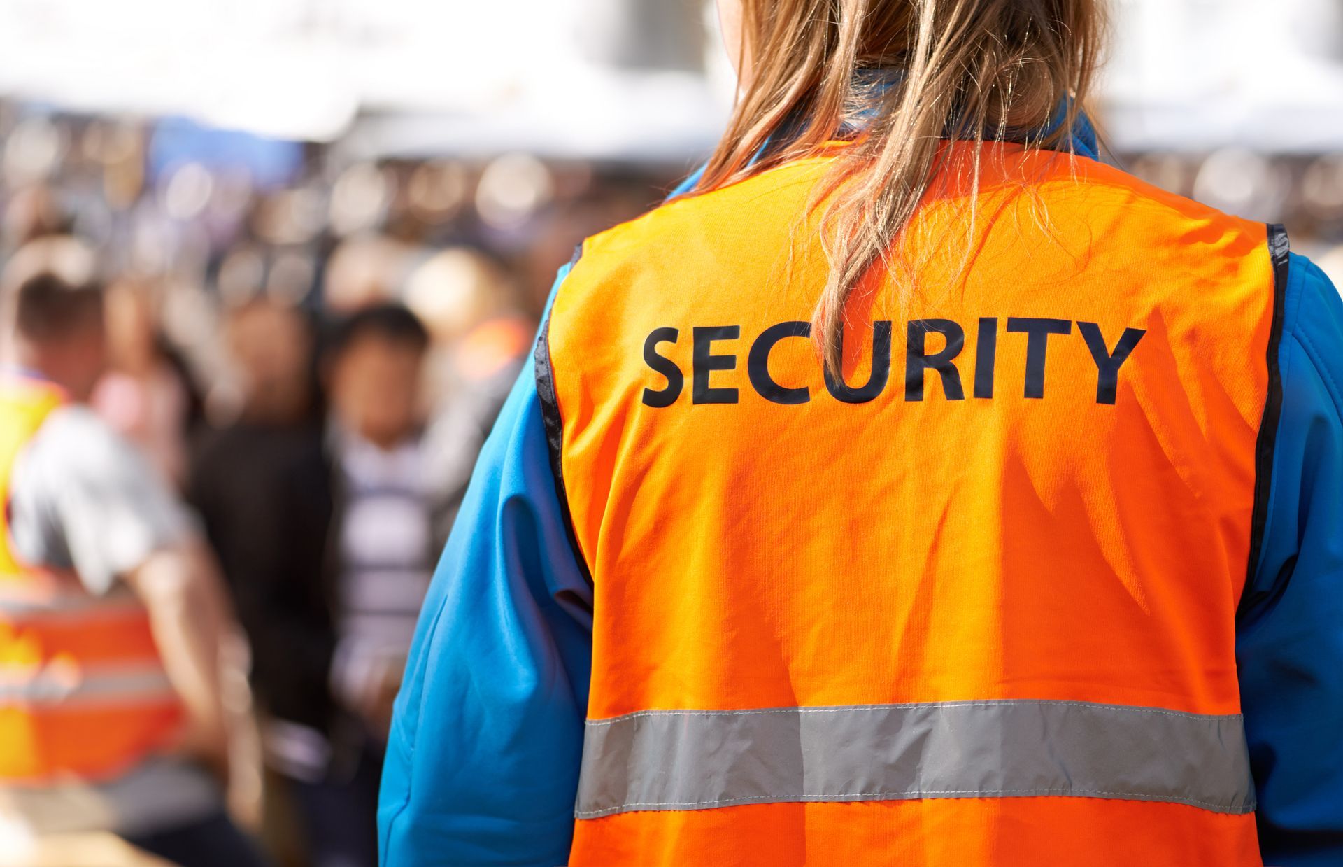 Security guard wearing orange vest, watching a crowd.