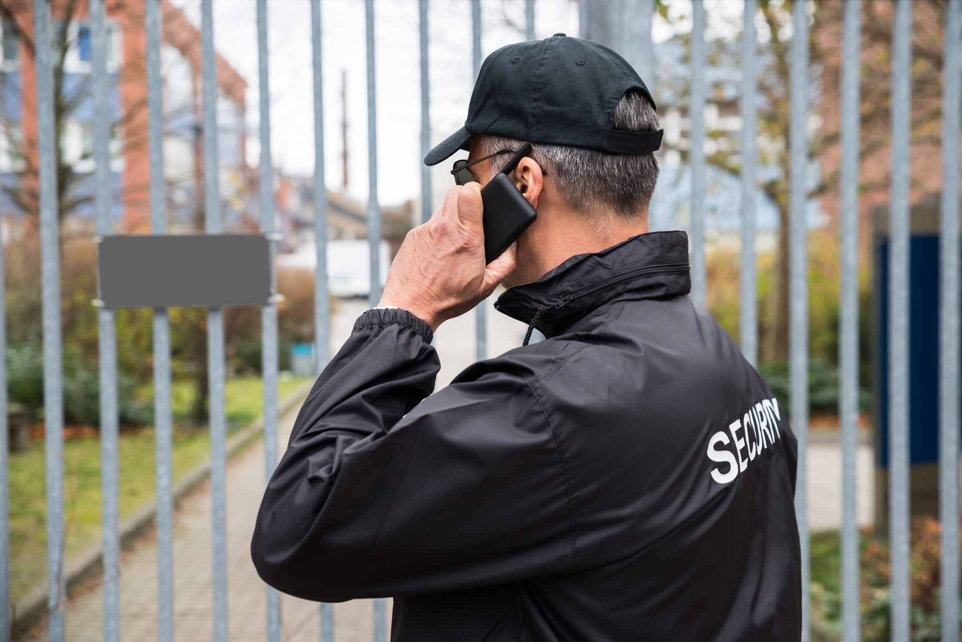 Security guard talking on a phone by a metal gate, wearing a black jacket and cap.