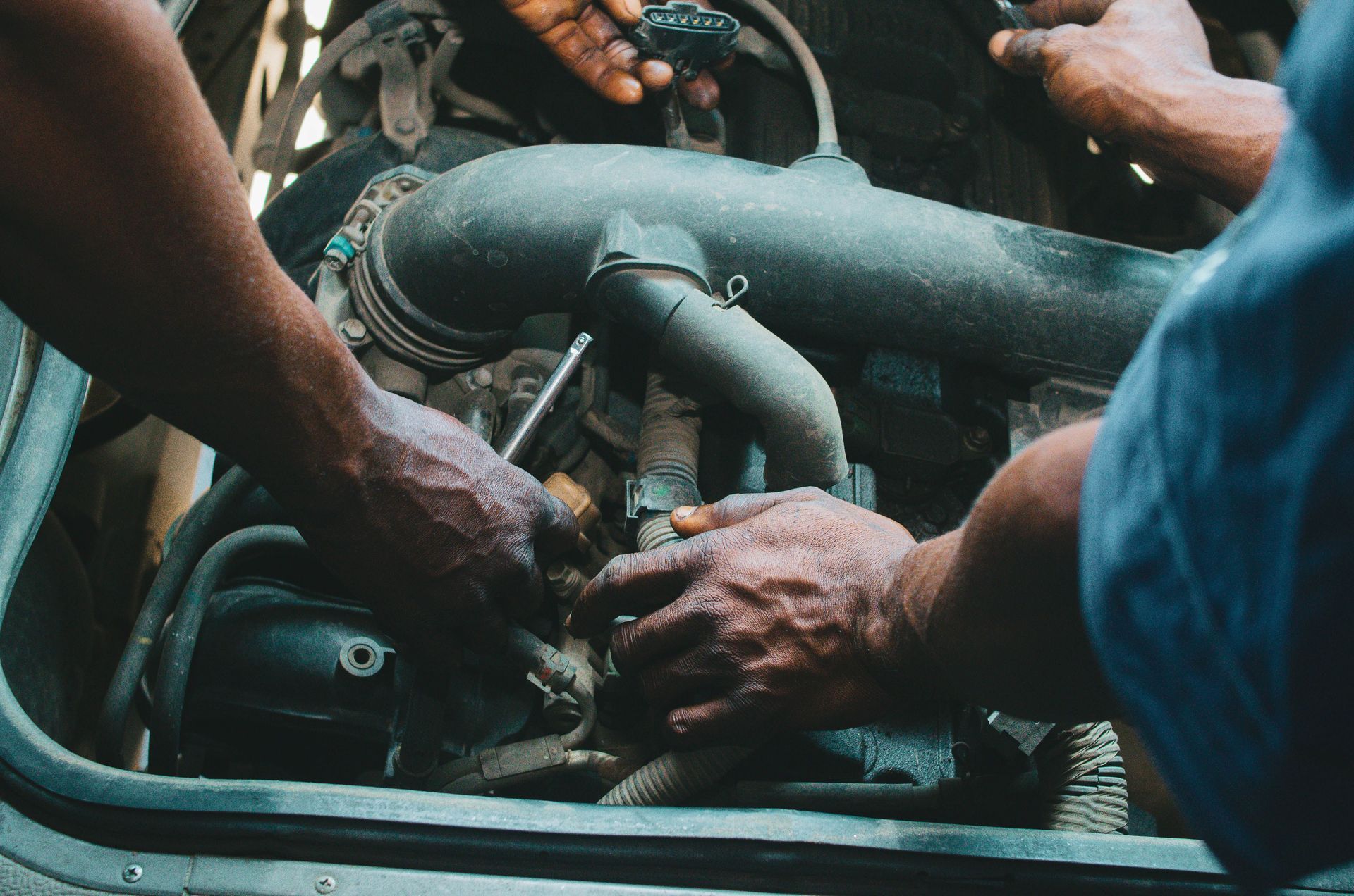 Hands working on a car engine in a crowded workshop, with tools and hoses visible.