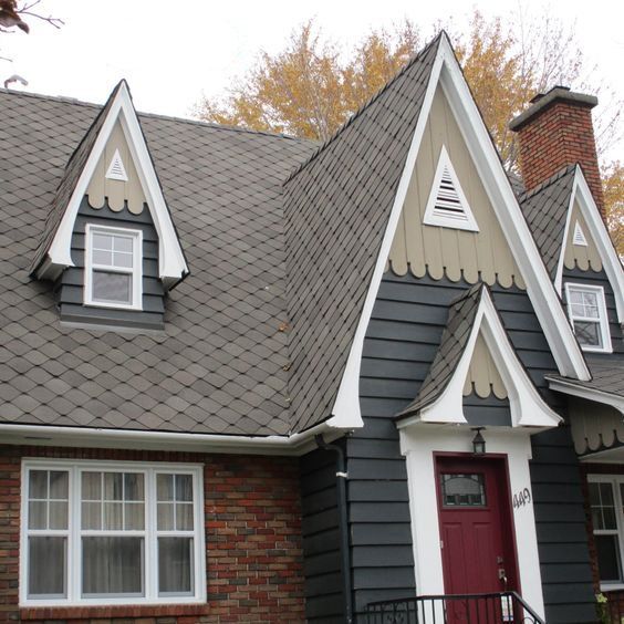Maison de style cottage avec revêtement gris foncé et porte rouge. Toiture marron, touches de brique et boiseries blanches.