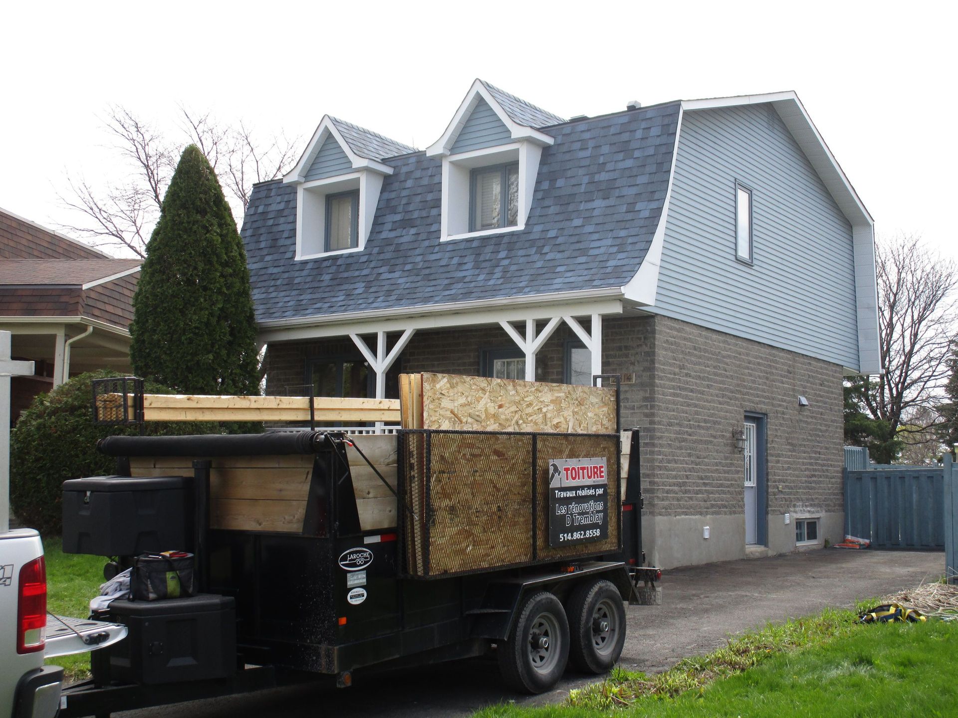 Une remorque chargée de bois devant une maison à deux étages avec un toit bleu et un extérieur en briques.