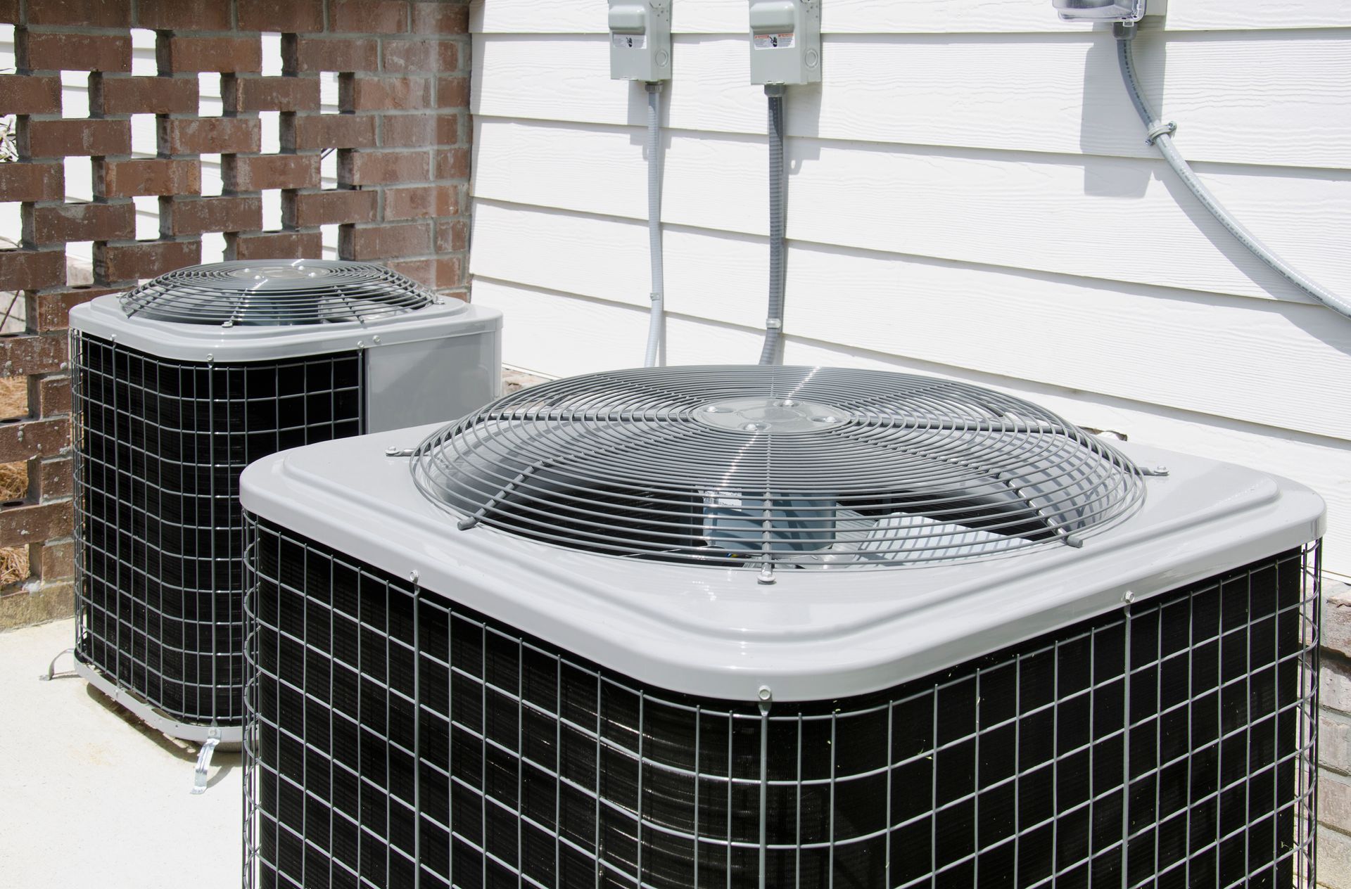 Two outdoor air conditioning units beside a house, with a brick wall and white siding behind them