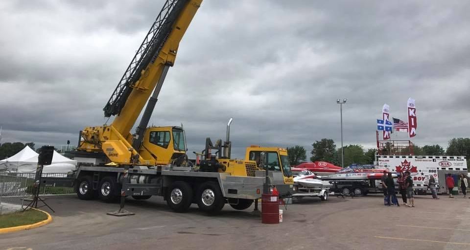 Grue jaune sur le plateau d'un camion lors d'un événement en plein air avec des véhicules d'urgence et des personnes ; ciel nuageux.