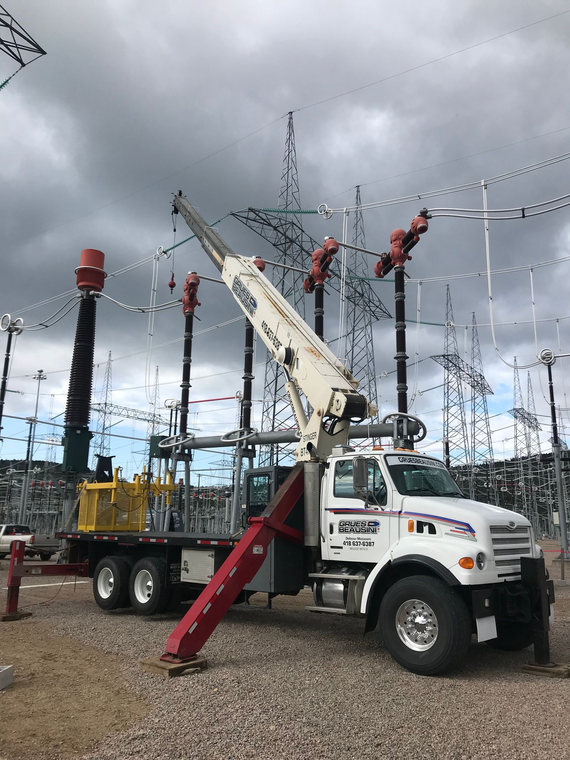 Un camion blanc équipé d'une grue dans un poste de transformation électrique, à proximité de lignes électriques.