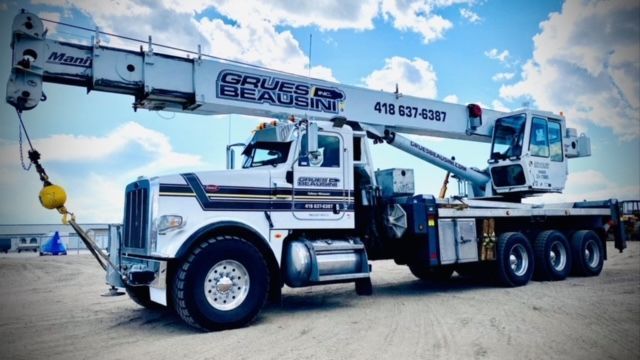 Camion-grue blanc, inscription « Grues Beausoleil », flèche déployée sous un ciel bleu.