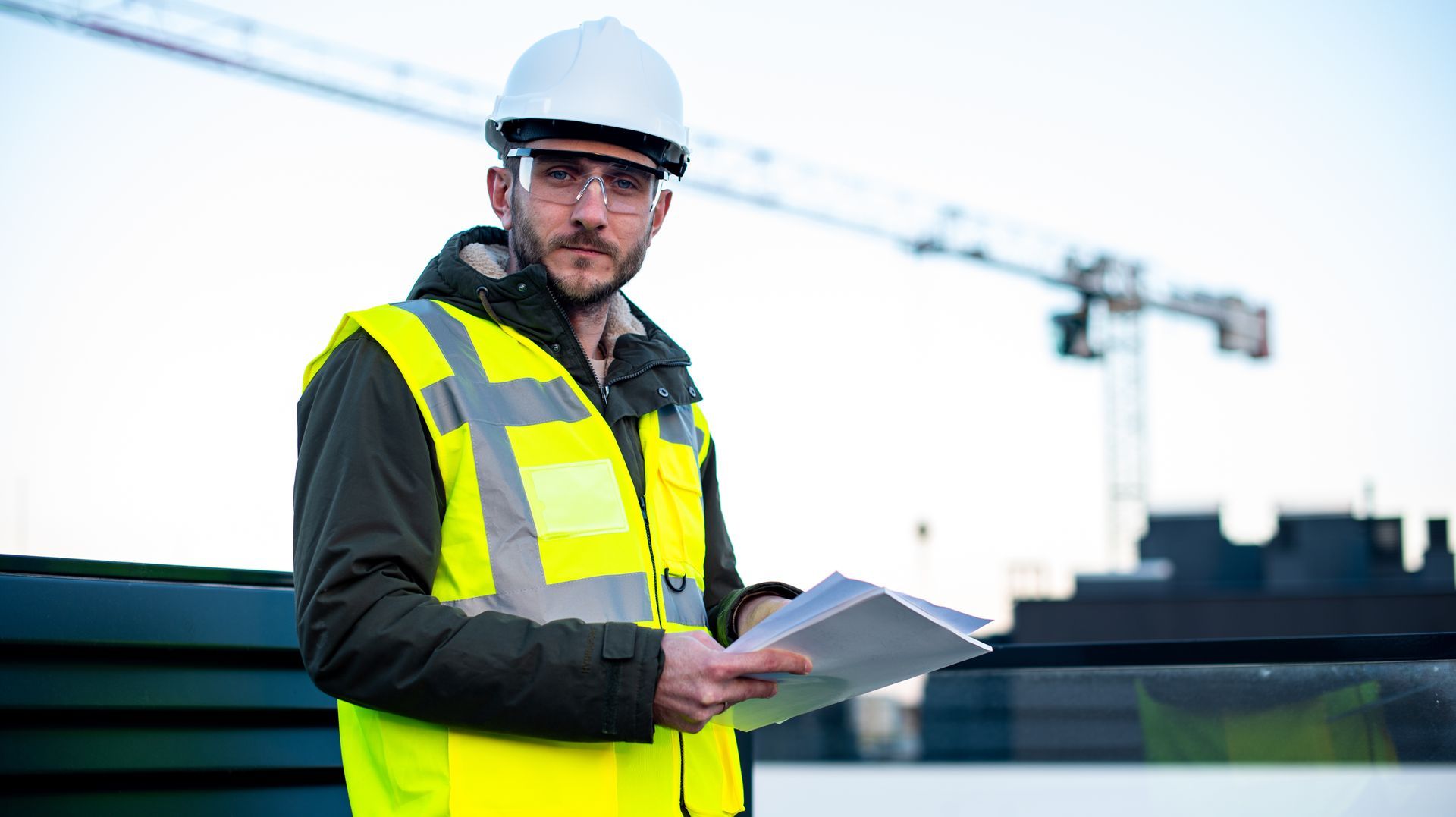 Un ouvrier du bâtiment, portant un casque et un gilet de sécurité, tient des papiers près d'une grue.
