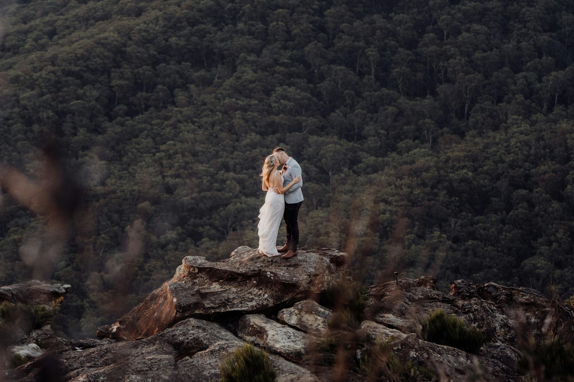 A bride and groom are kissing on top of a mountain.