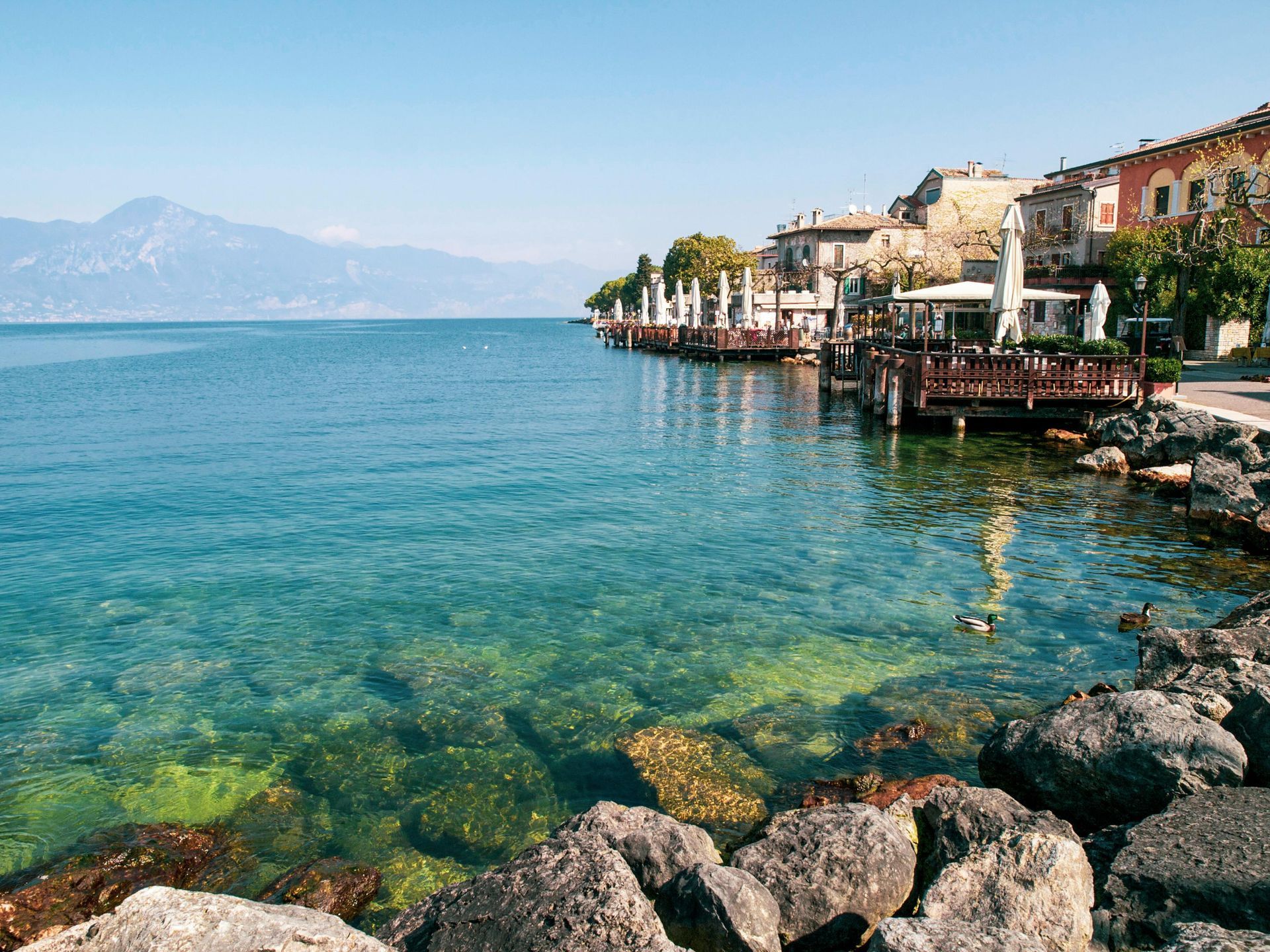 Lakeside view with clear turquoise water, rocky shore, and buildings along the coast on a sunny day.