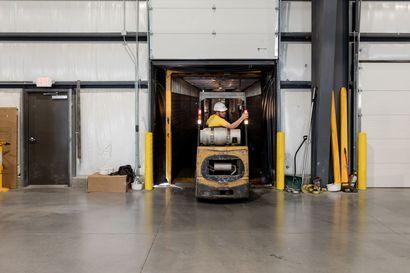 Forklift operator in a warehouse doorway, entering a dock. The operator wears a hardhat and a yellow shirt. The setting is a gray warehouse. Forklift operator in a warehouse doorway, entering a dock. The operator wears a hardhat and a yellow shirt. The setting is a gray warehouse.