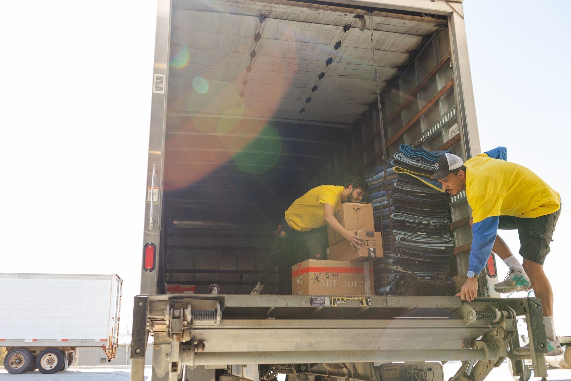 Two people loading boxes into a truck. One on the liftgate, one inside. Bright yellow shirts, sunny outdoor setting. Two people loading boxes into a truck. One on the liftgate, one inside. Bright yellow shirts, sunny outdoor setting.