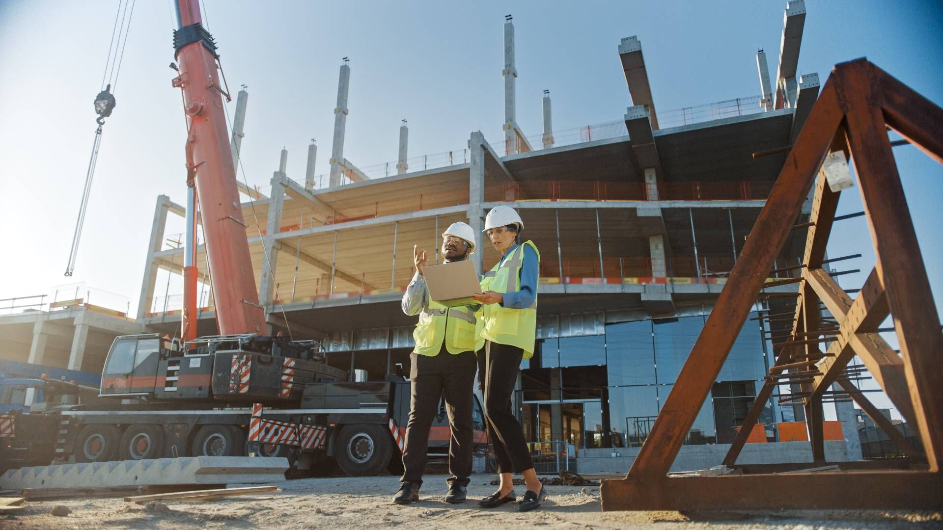 Two construction workers in safety vests and hard hats reviewing a tablet at a building site with a crane.