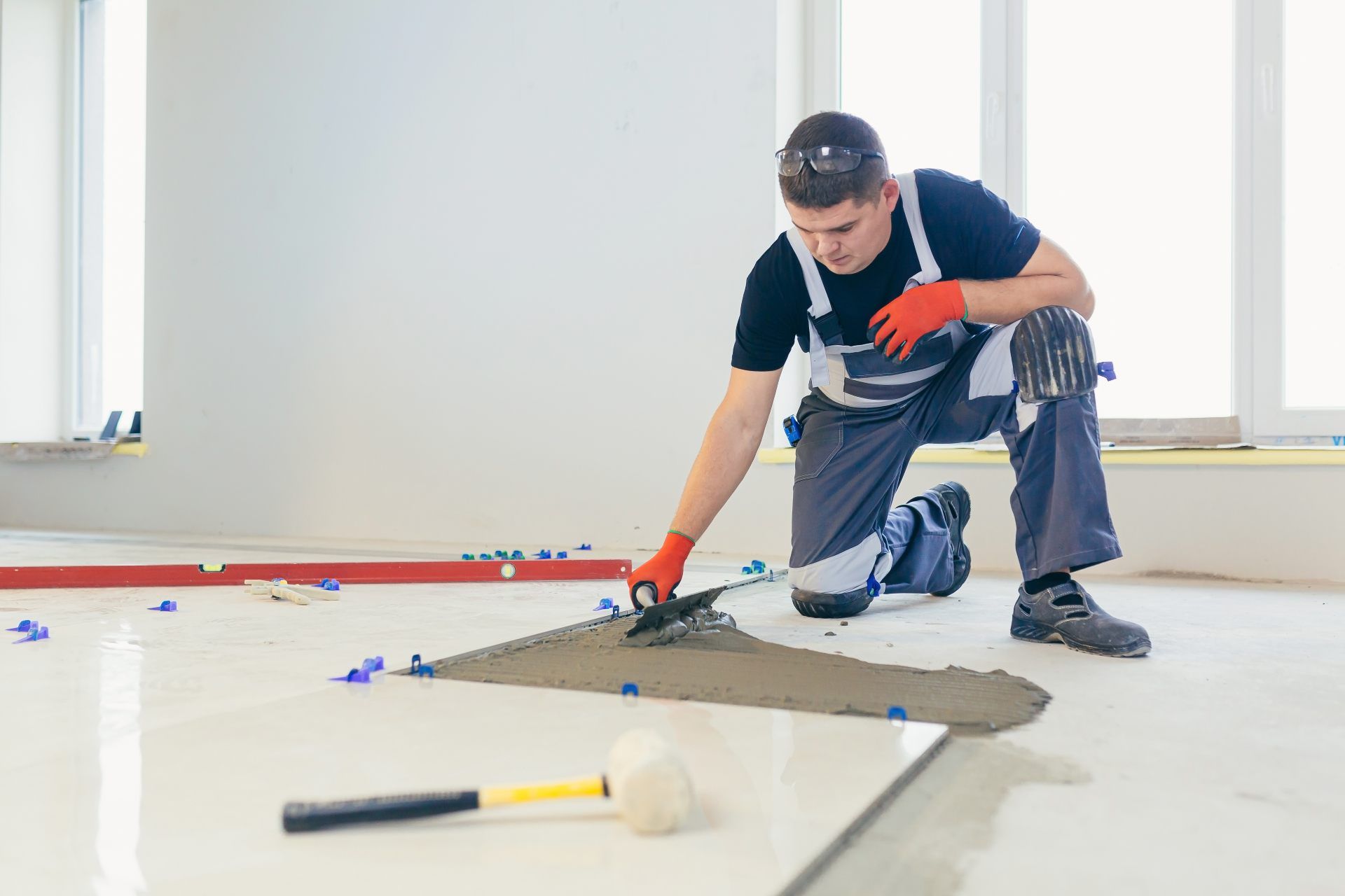 Man kneeling, applying mortar to tile floor with a trowel, surrounded by tools and tiles in a bright room.