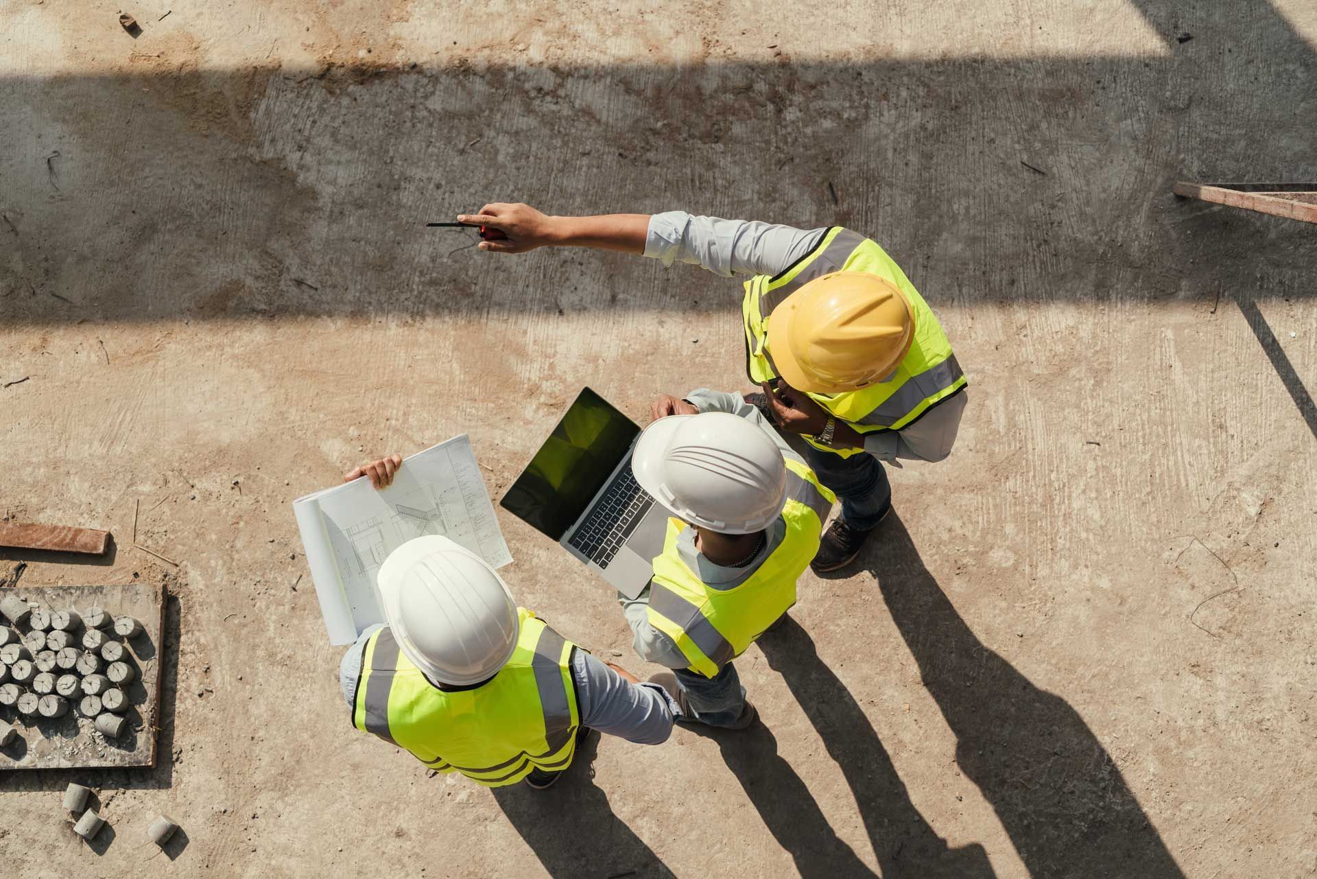 Construction workers in safety vests and hard hats reviewing blueprints and laptop on site.
