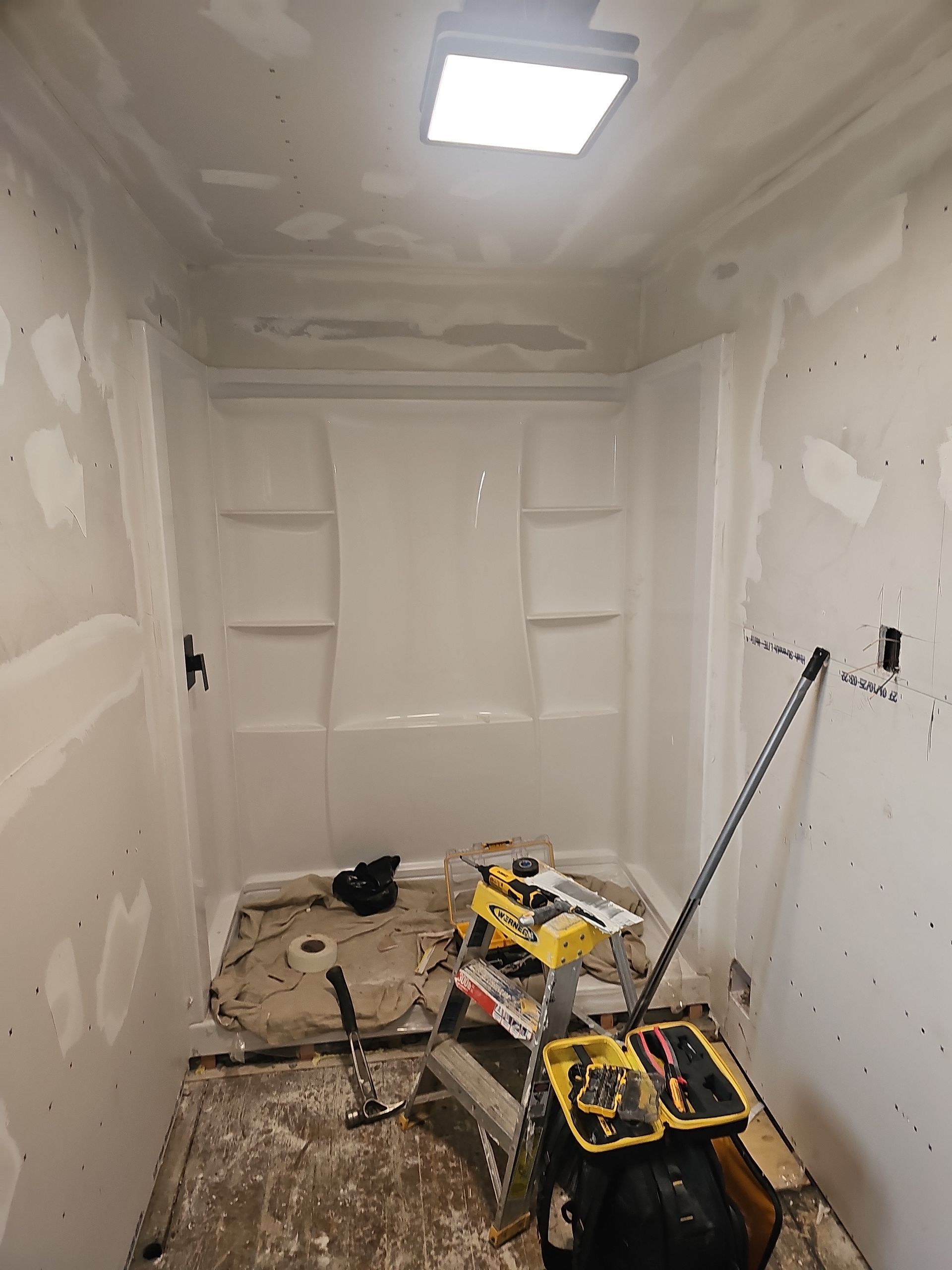 Bathroom under construction, white shower stall, drywall, tools, and ceiling light.