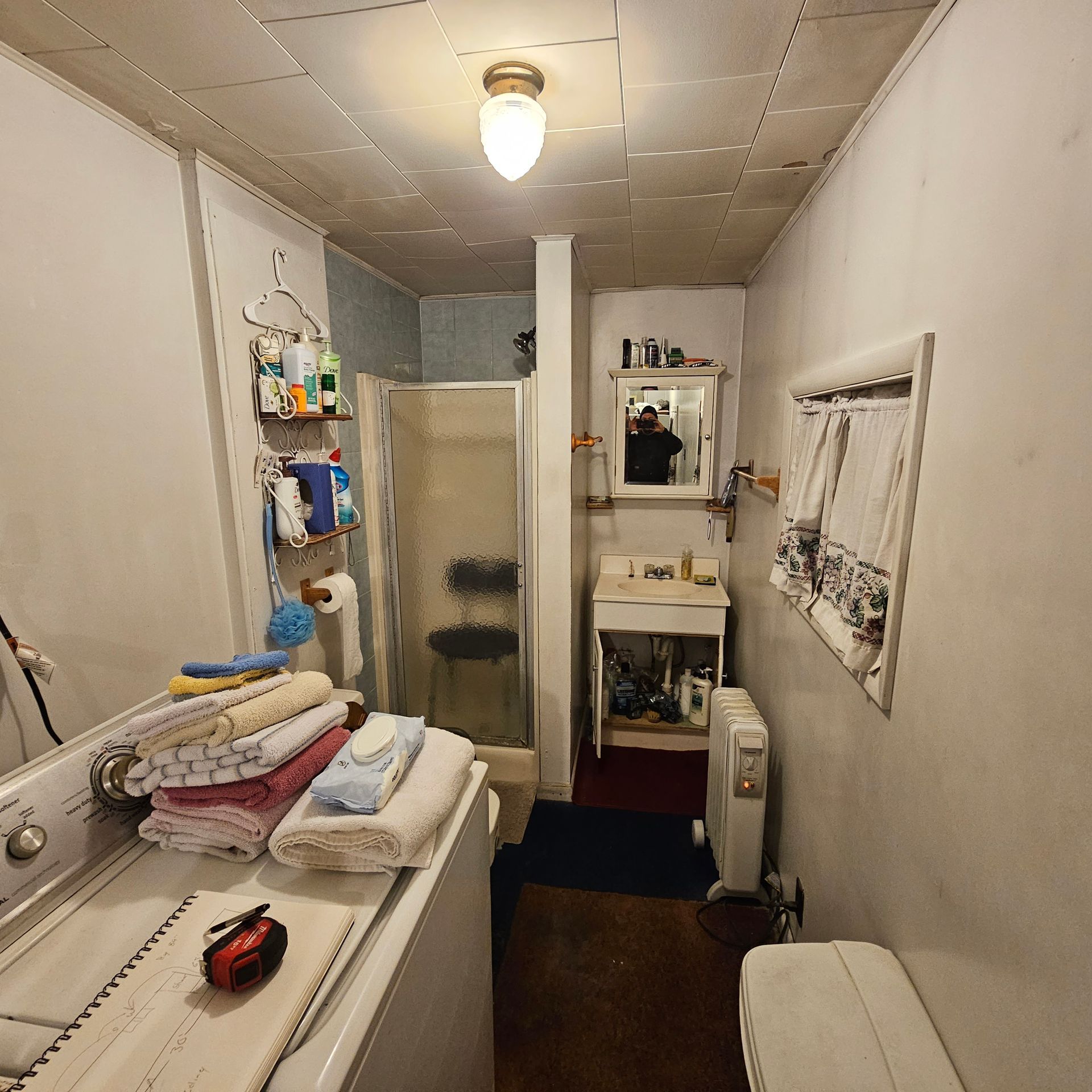 Small, cluttered bathroom with washing machine, shower, sink, and shelves with toiletries.