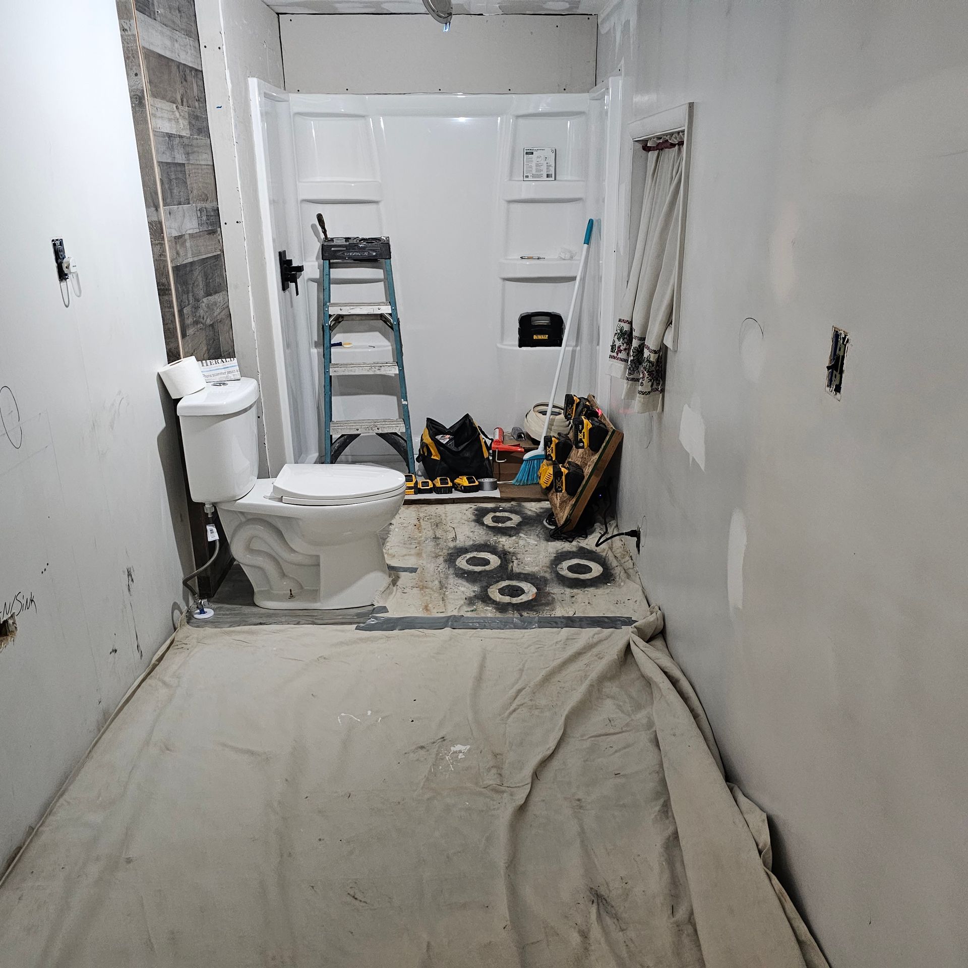 Bathroom under renovation with toilet, shower, ladder, and tools on drop cloth.