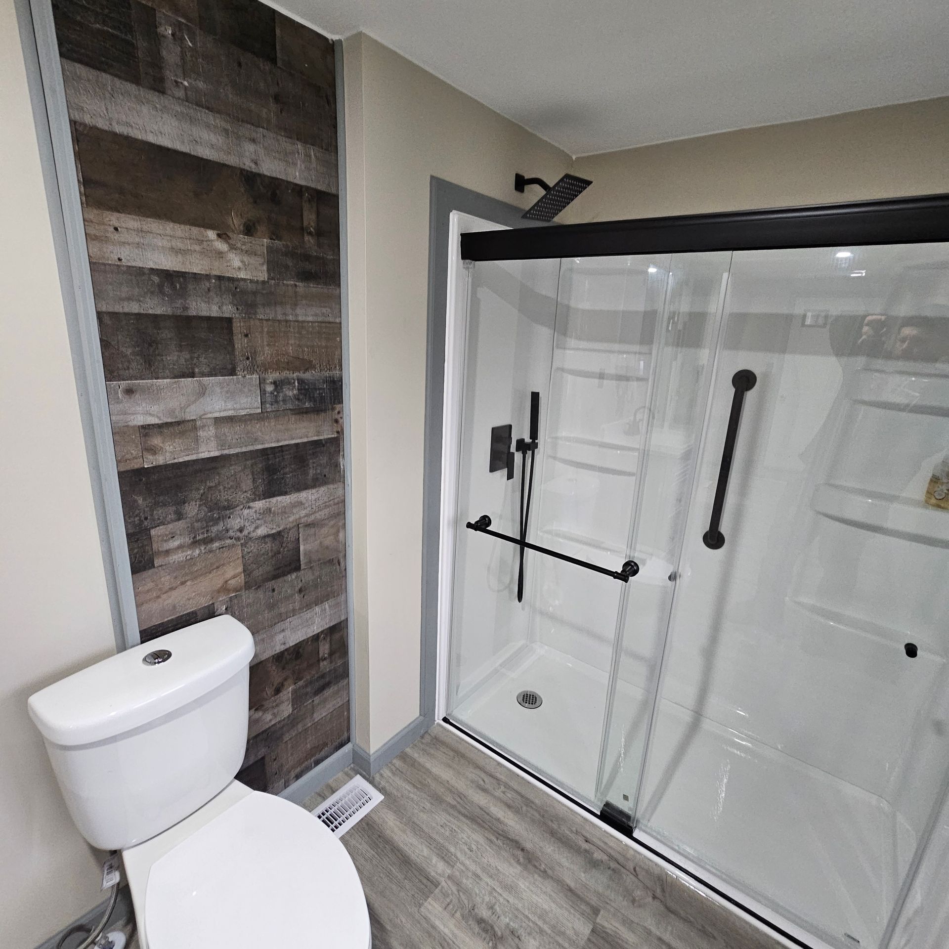 Bathroom with white toilet, wood-paneled wall, and a black-framed shower with grab bars.