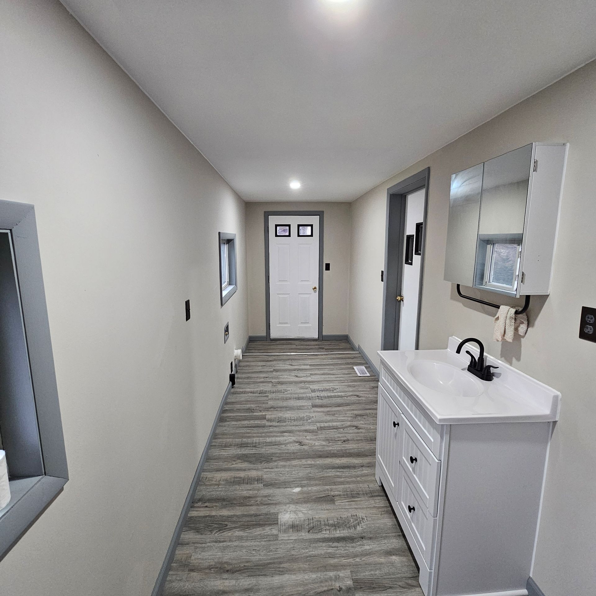 Long hallway with white walls, wood-look flooring, and a white vanity with black faucet.