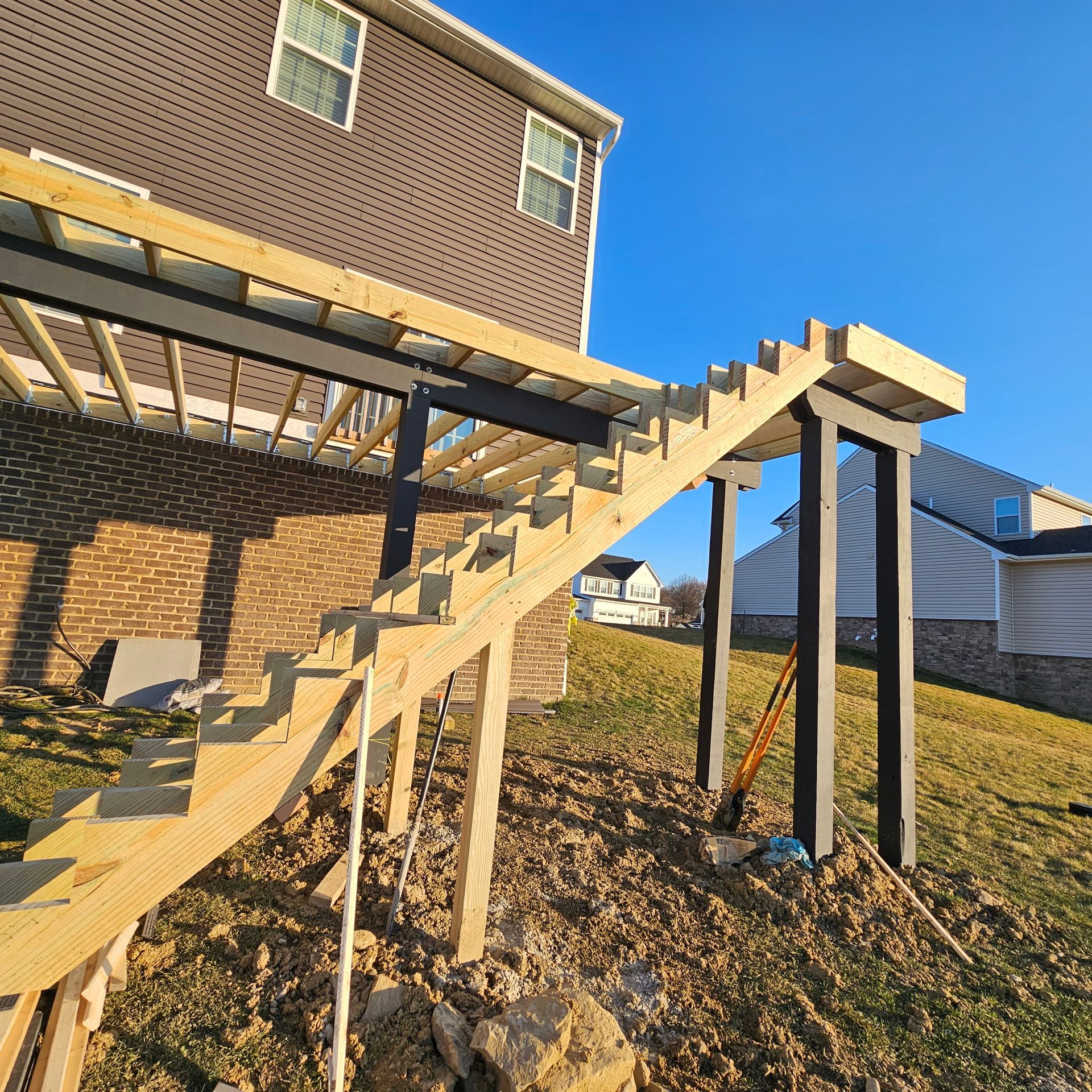 Wooden deck stairs under construction, attached to a house with a brick wall and a sloping yard.