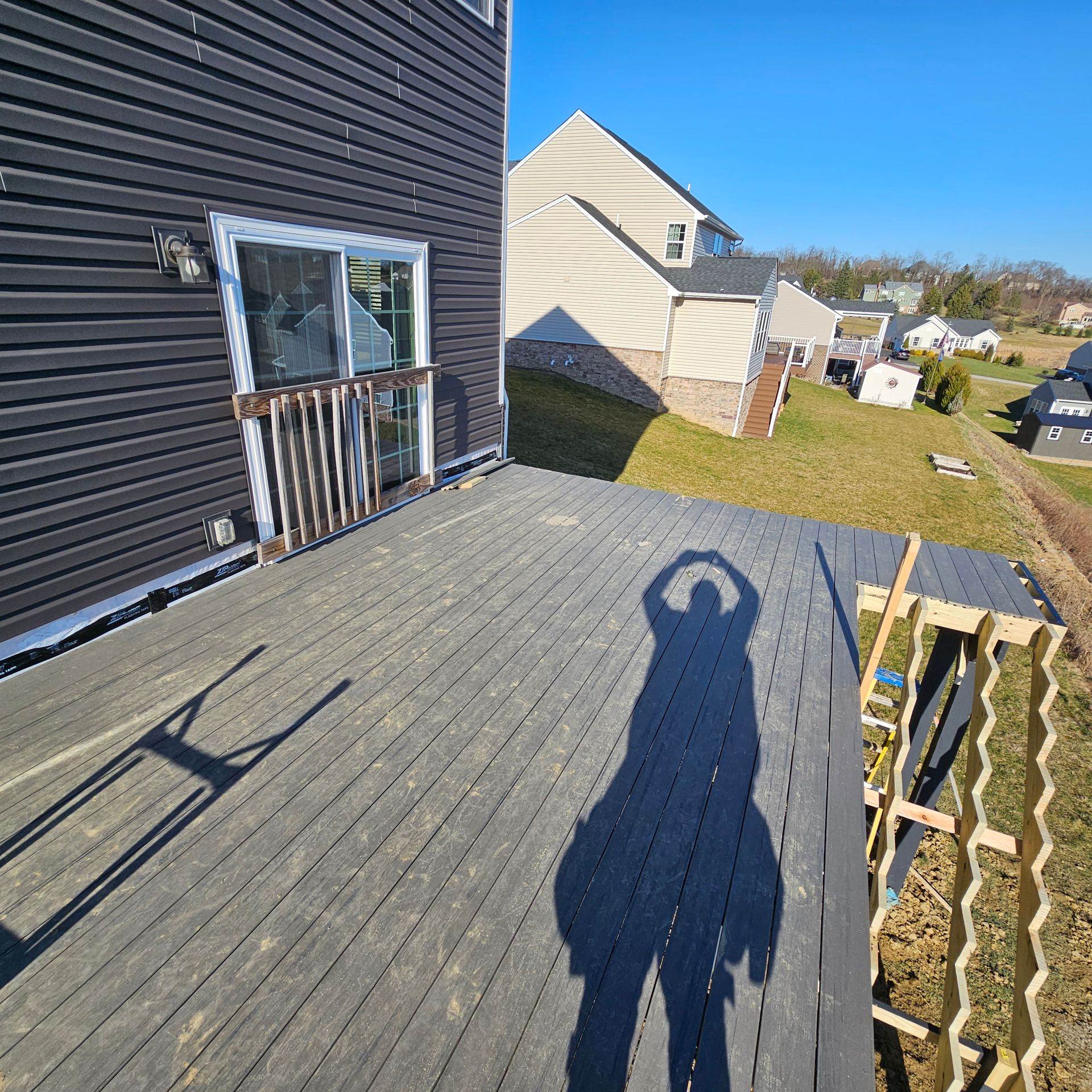 A person's shadow on a weathered gray deck near a house; blue sky background.