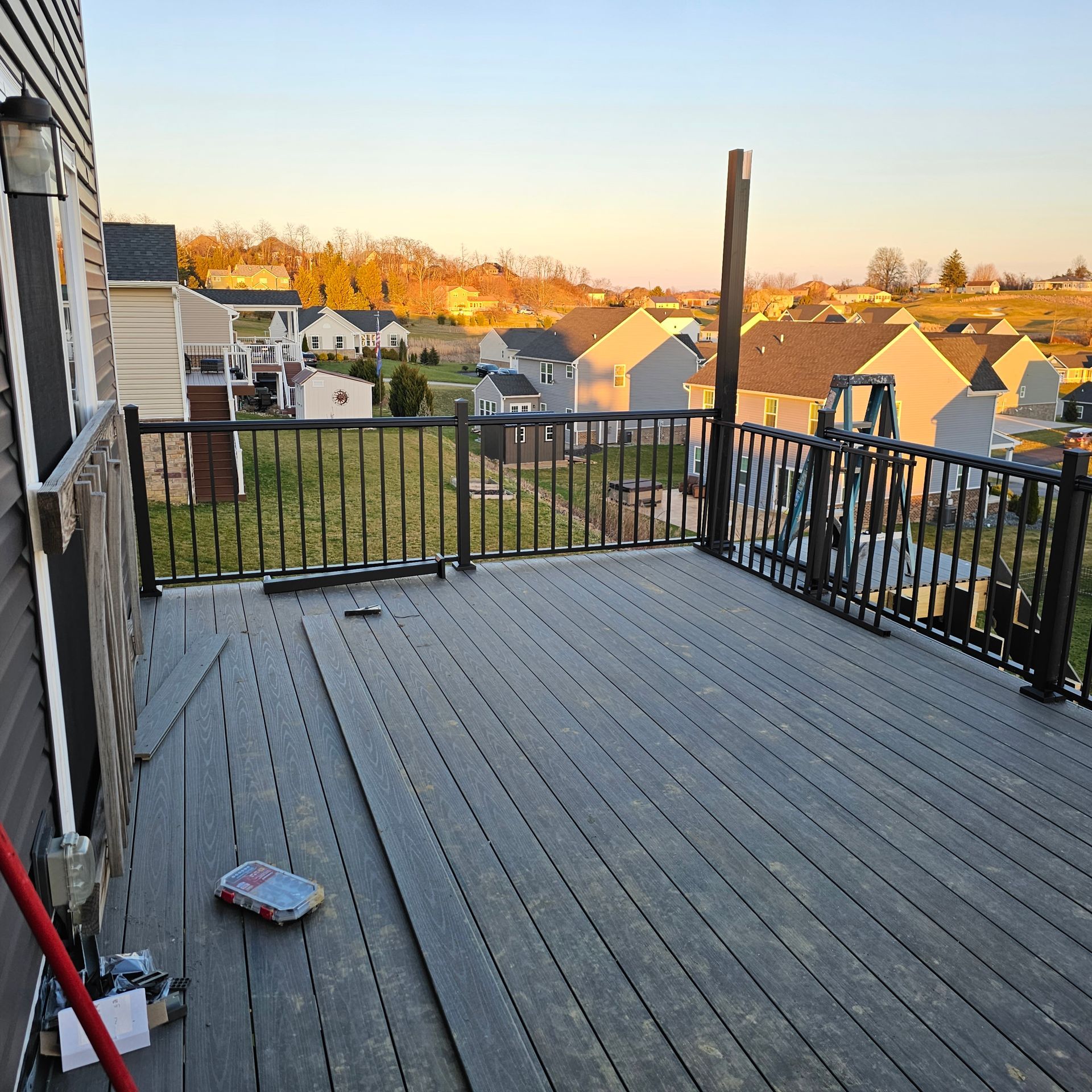 A composite deck with black railing overlooks a suburban landscape at sunset. Construction tools are visible.