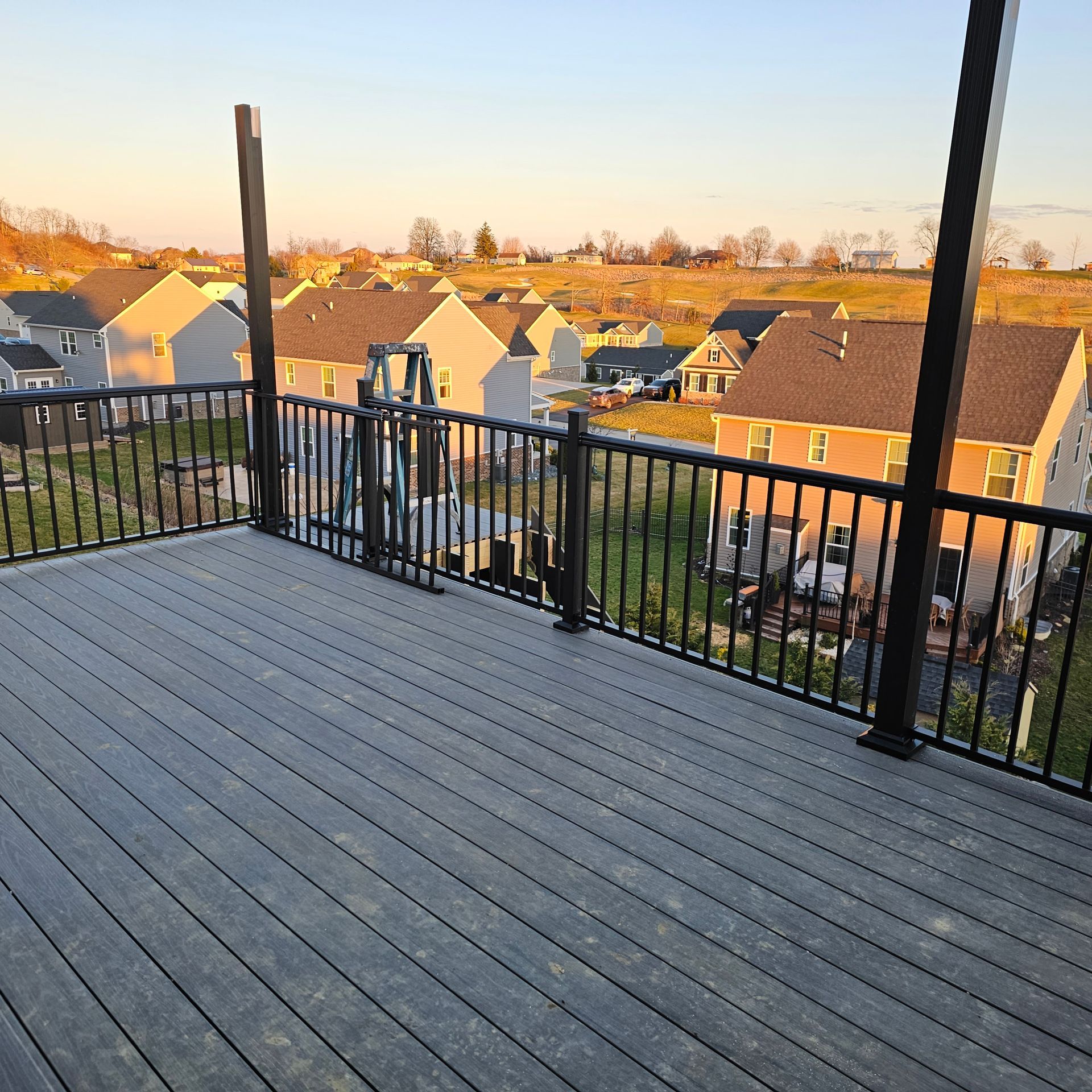 Deck with black railing, overlooking a suburban neighborhood at sunset. Gray composite decking.