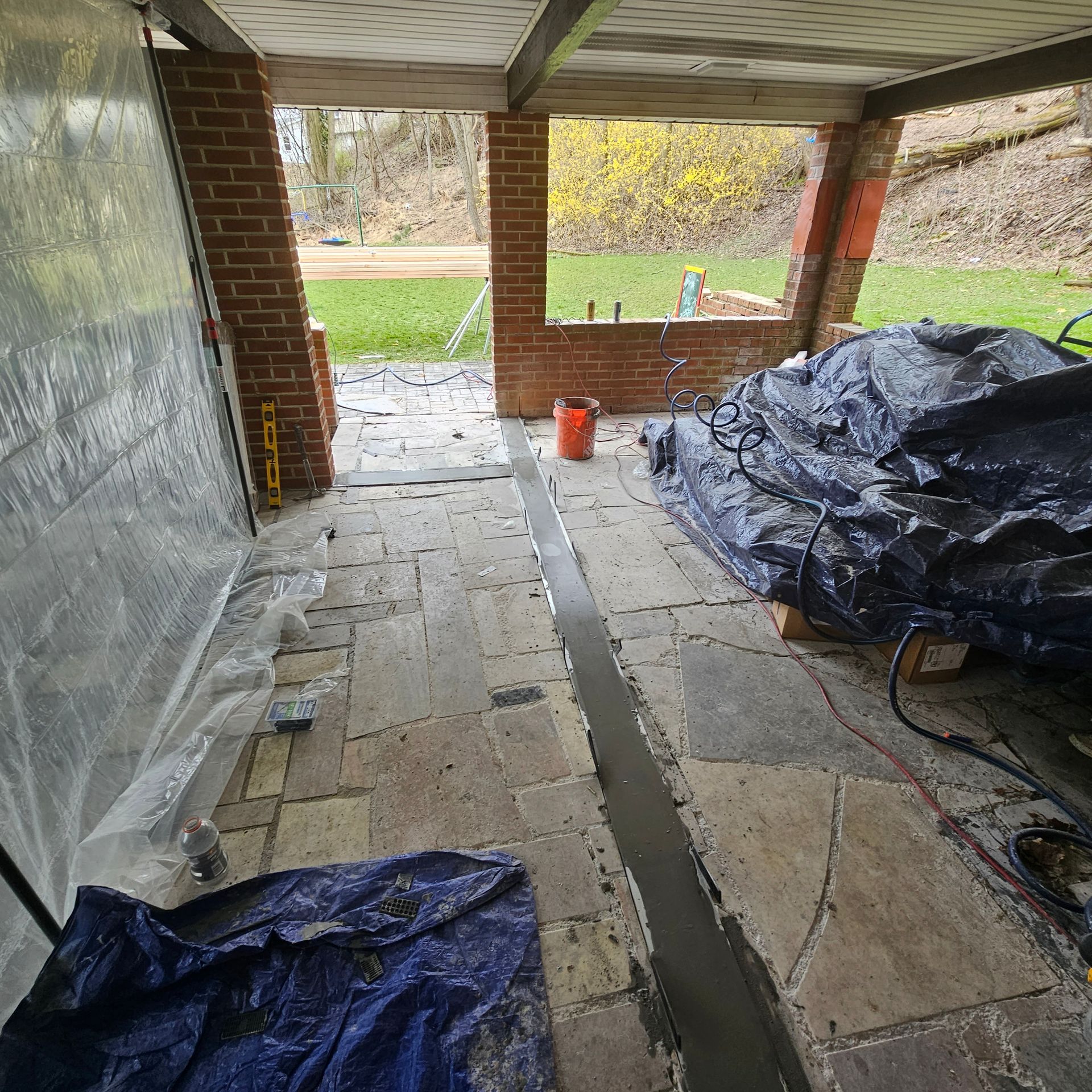 A brick-walled porch area under construction; concrete channel visible in the stone flooring, with tarps and tools nearby.