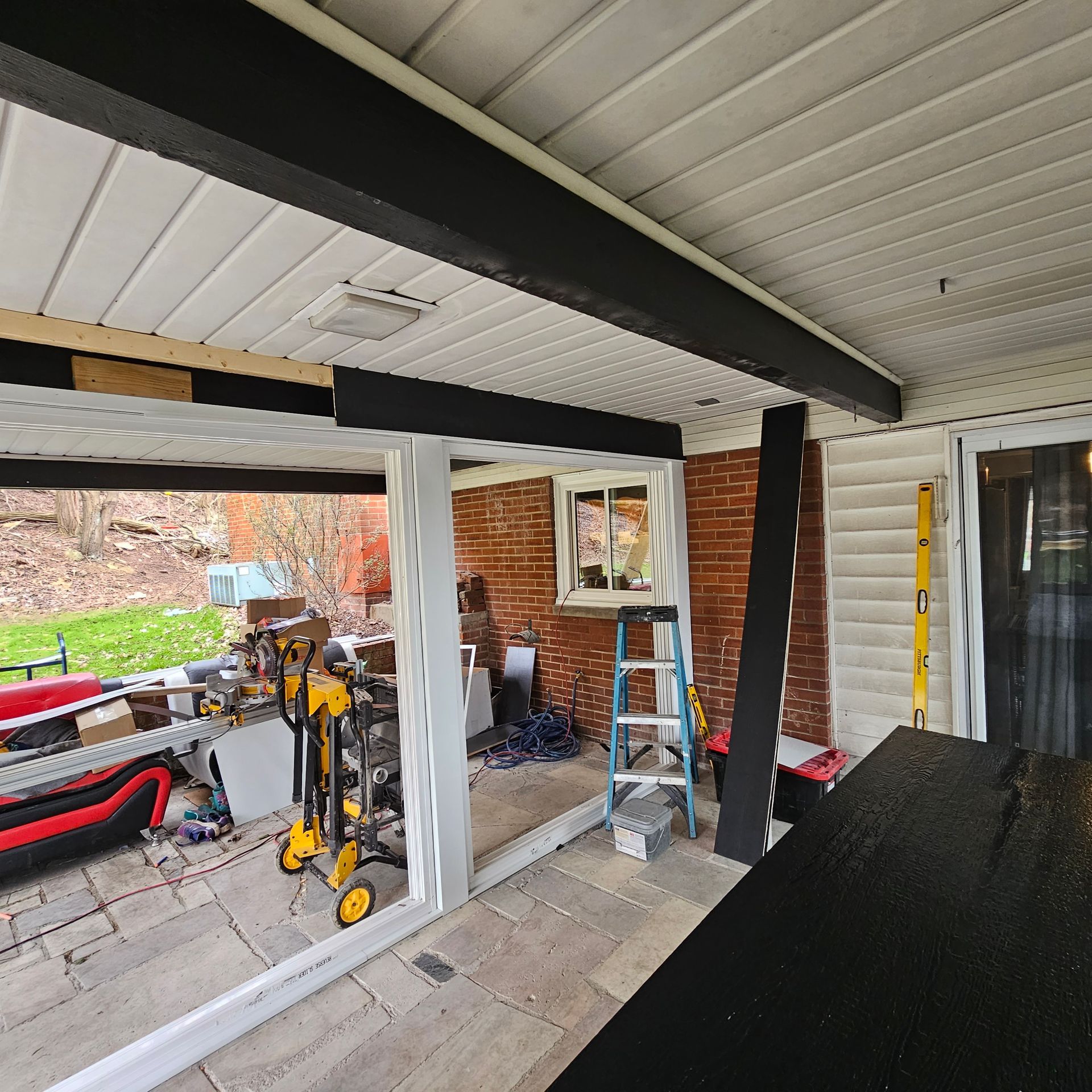Construction site: A patio roof with black beams, white paneling, tools, and brick wall.