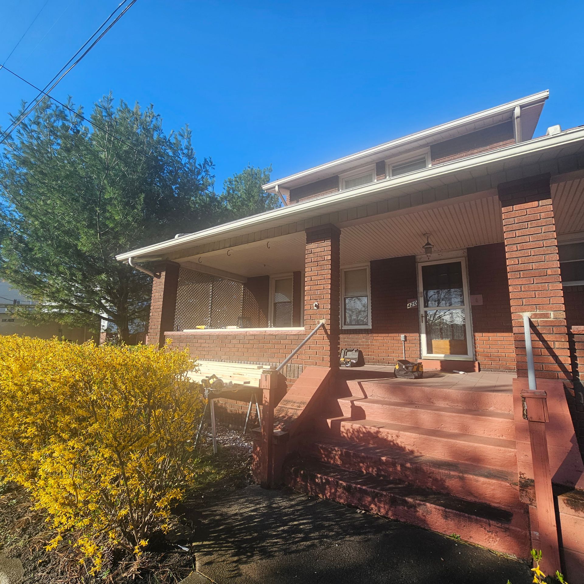 Brick house with covered porch, steps, and yellow flowering bush against a blue sky.