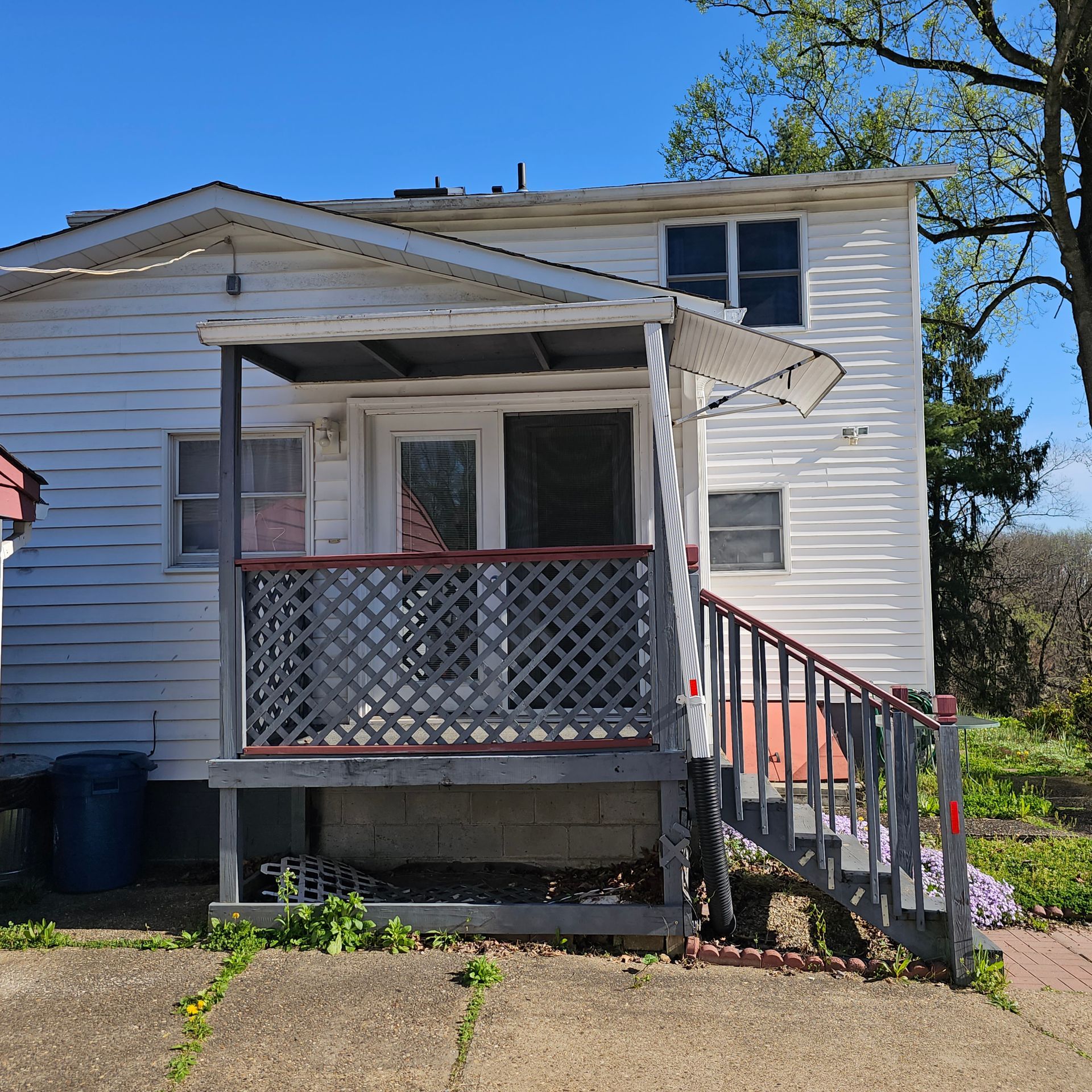 White house with porch, gray lattice railing, and red steps. Blue trash can and overgrown plants.