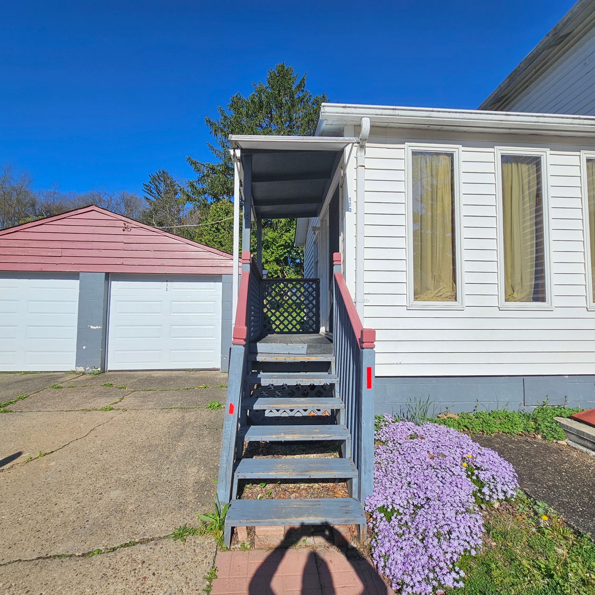 Exterior of a white house with a porch and steps, next to a garage; purple flowers in foreground.