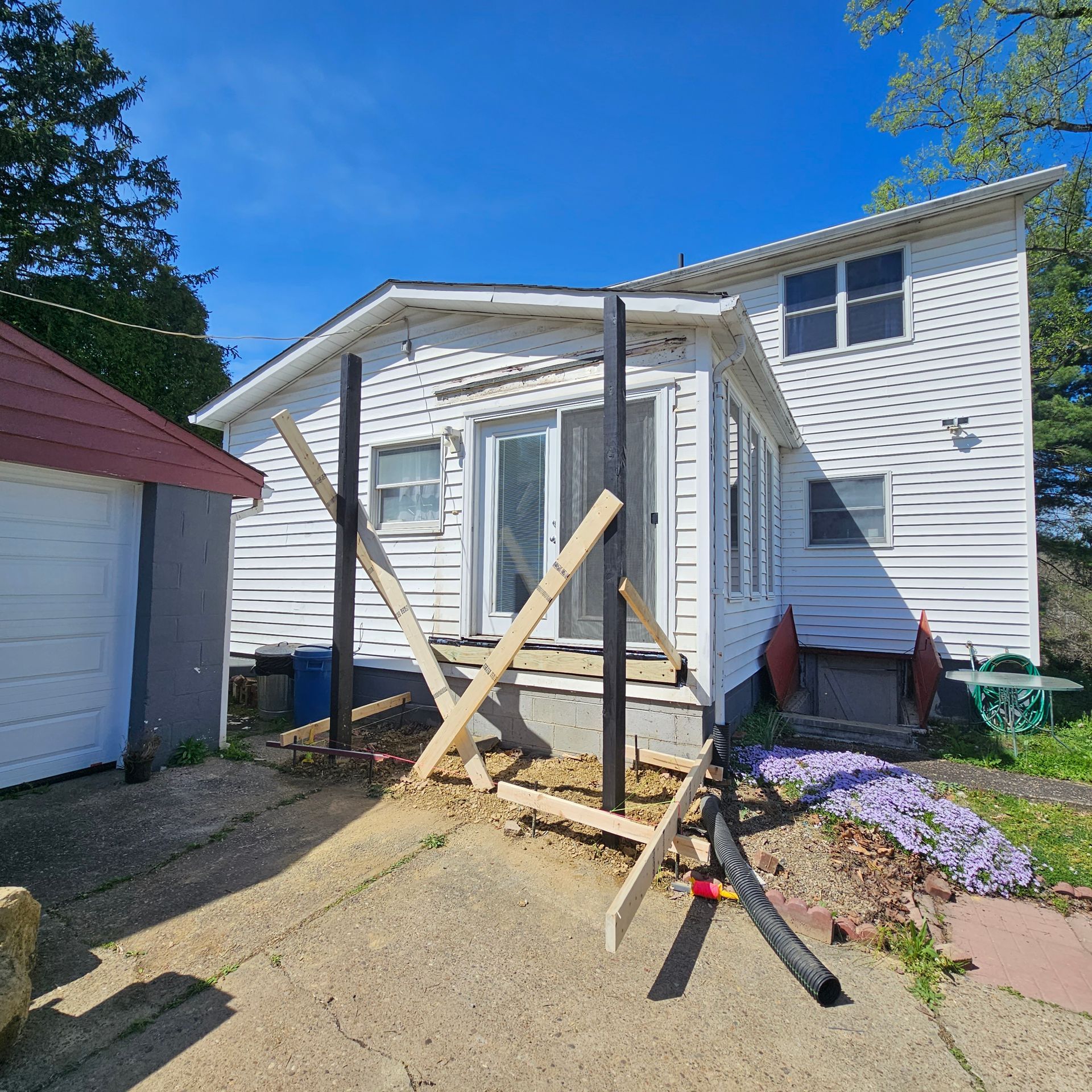 A white house with black posts and temporary wooden supports under a porch, on a sunny day.