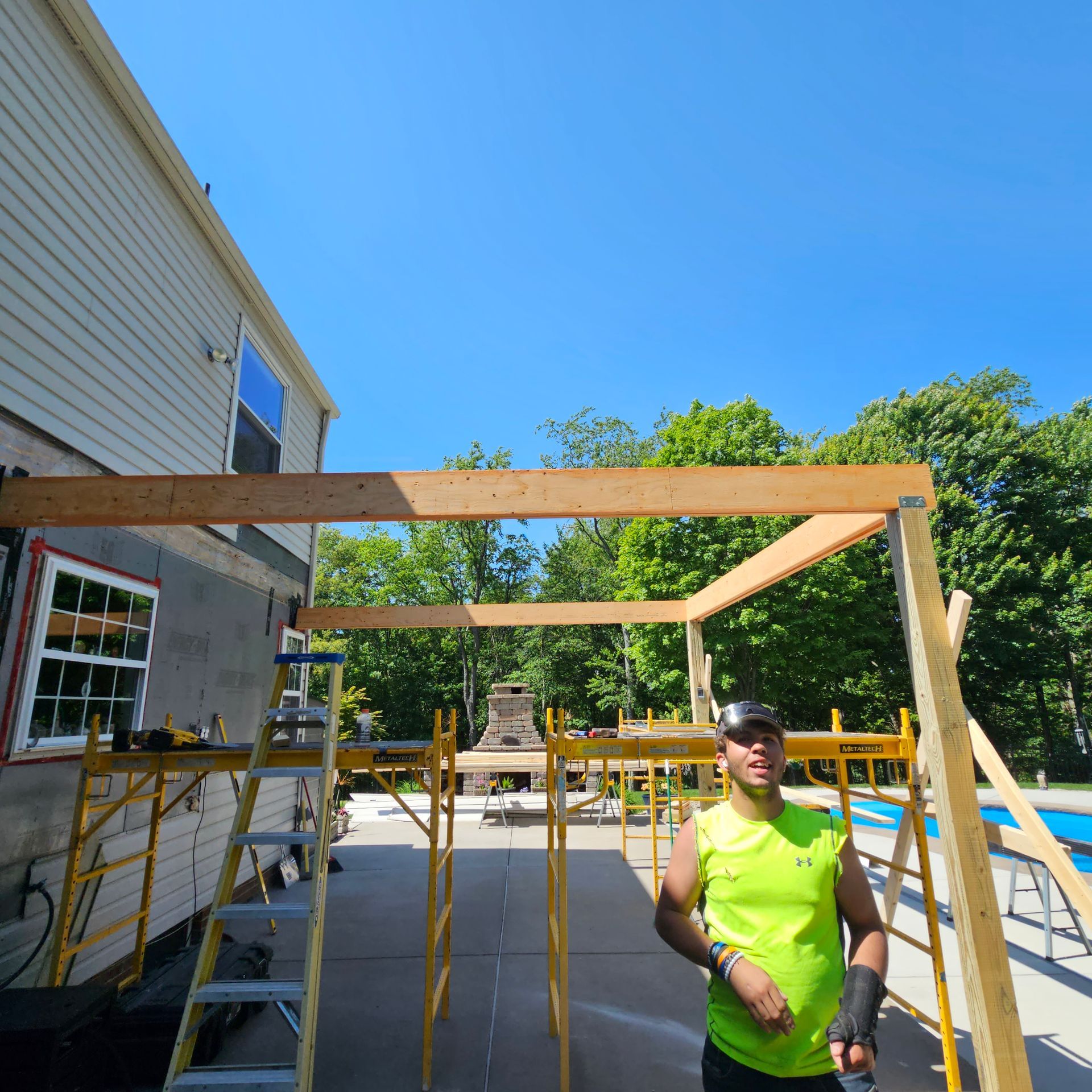 Man in neon green shirt building wooden structure near a house. Scaffolding, bright blue sky.