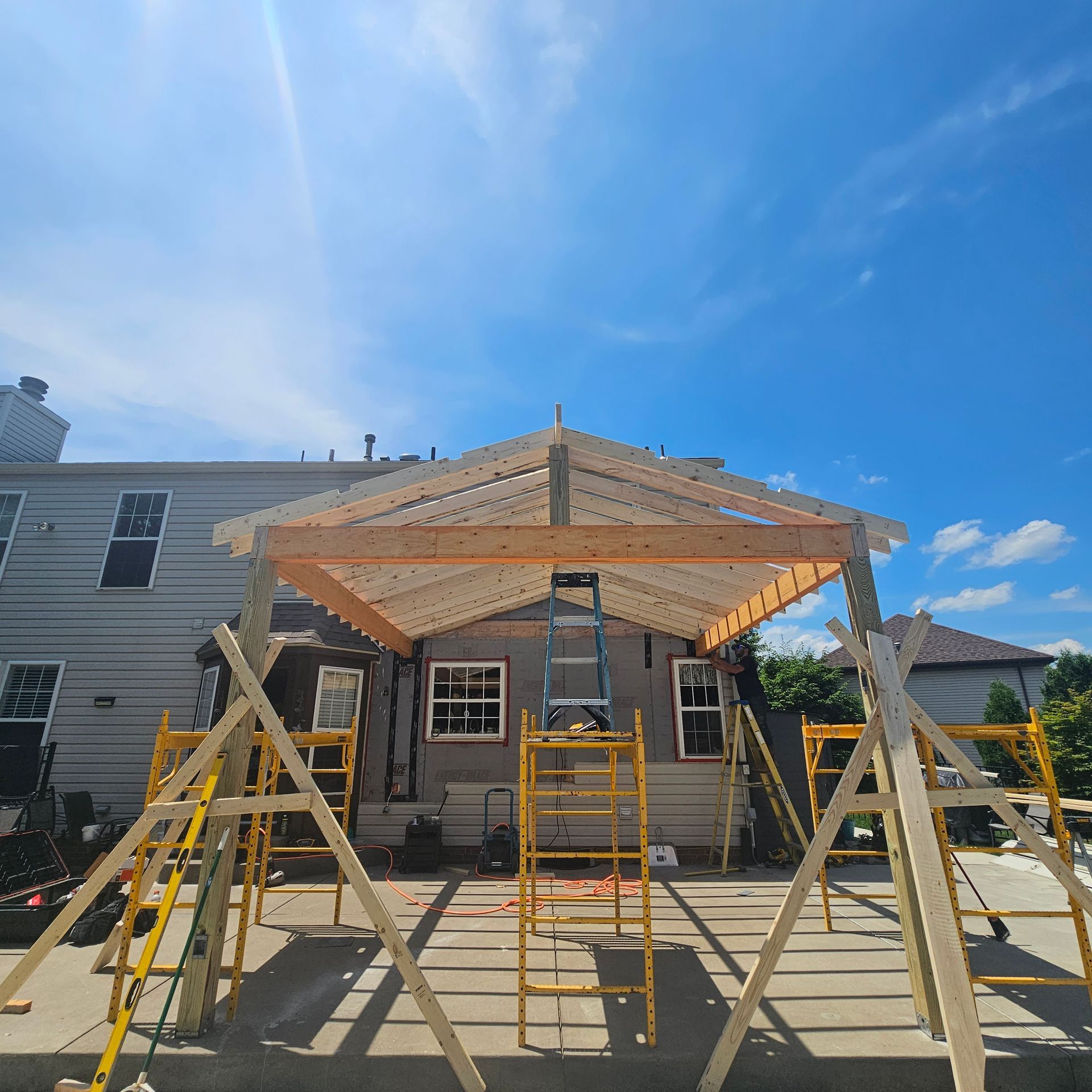 Wooden frame of a gazebo being built on a concrete patio, against the backdrop of a two-story house under a blue sky.