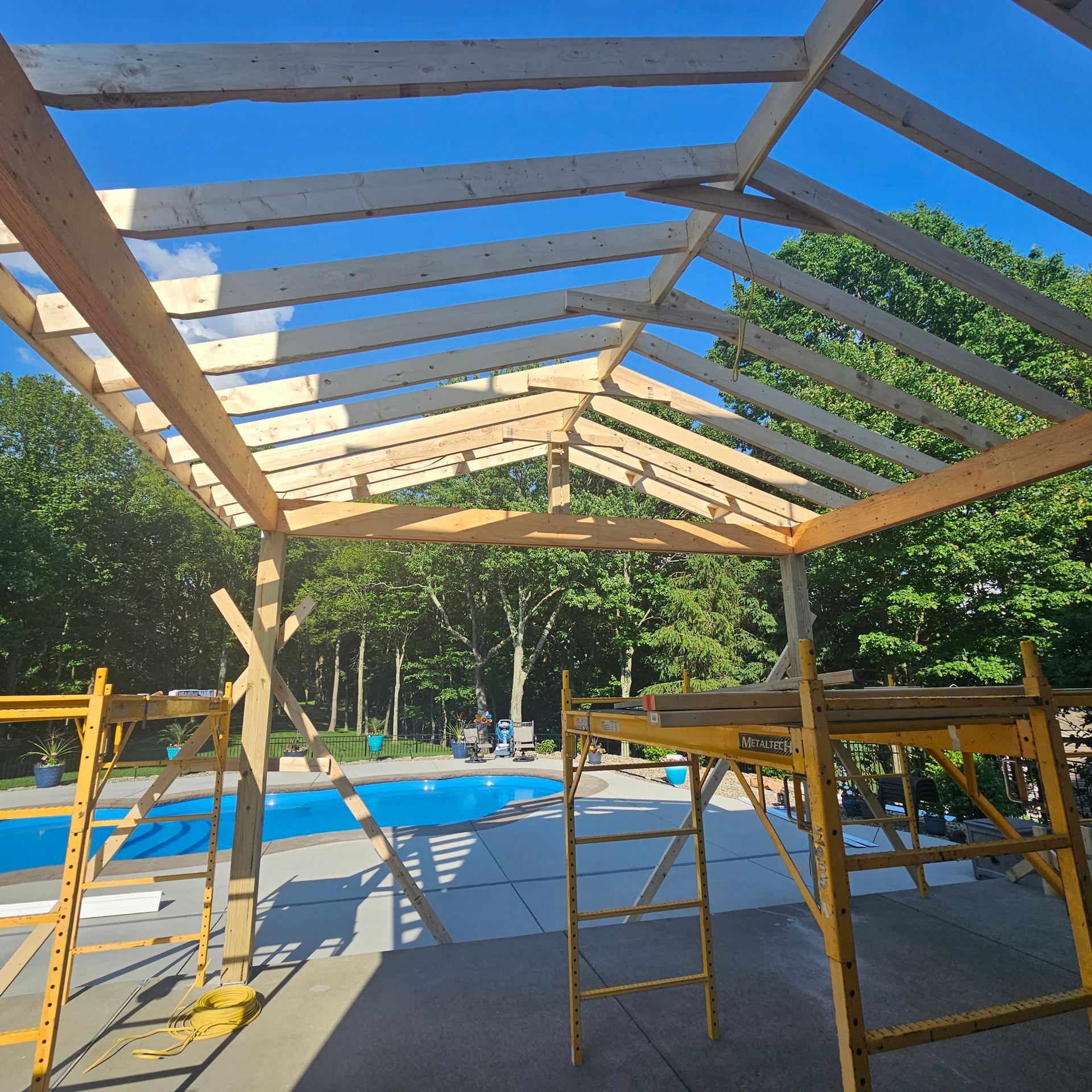 Wooden frame pavilion under construction near a pool. Sunny day, blue sky. Scaffolding visible.