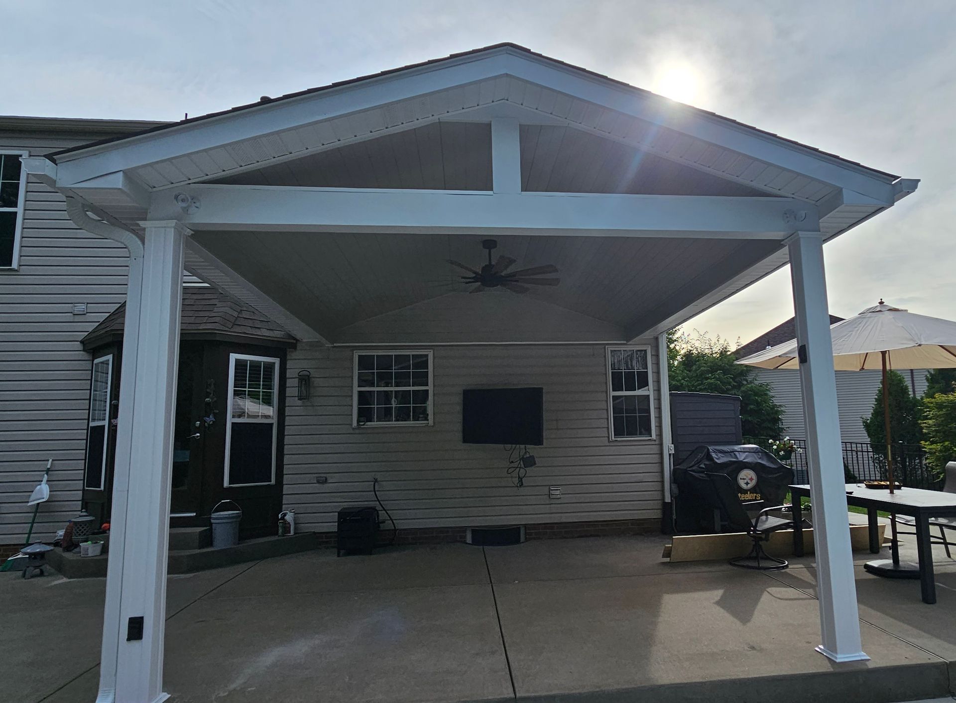 Covered patio with a white roof, ceiling fan, and concrete floor, adjacent to a house.