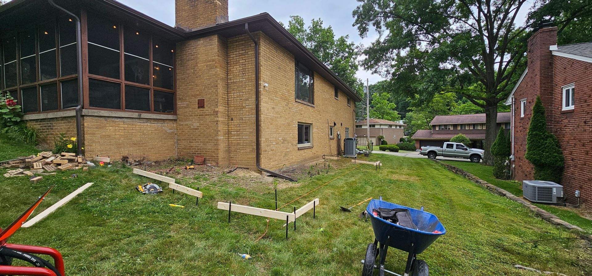 A brick house with a screened-in porch and a grassy yard. A blue wheelbarrow and a truck are visible.
