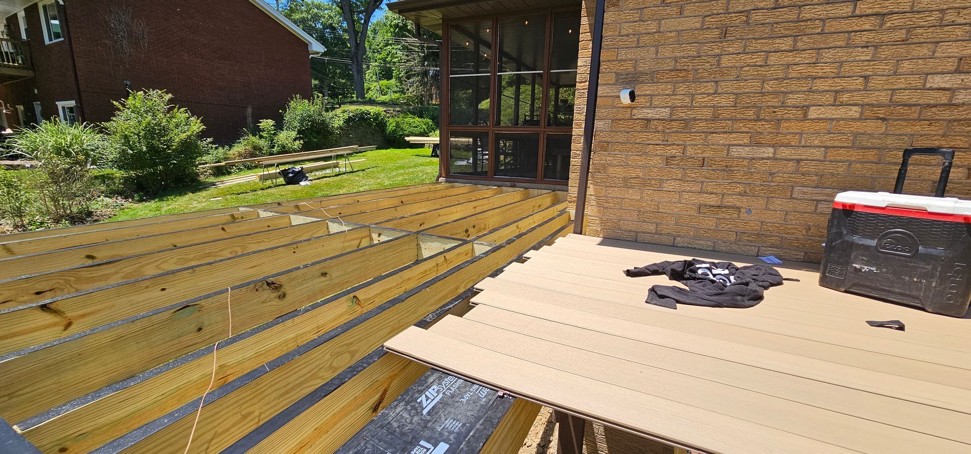 Deck construction in progress; wooden boards being laid, brick wall, screened porch, lush green yard.