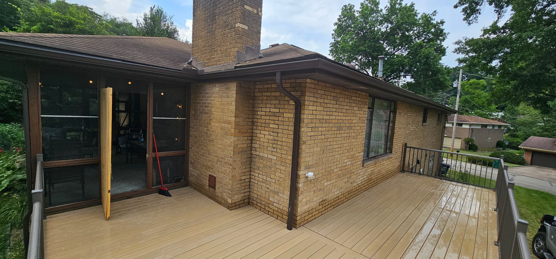 A house with brick siding, a deck, and trees. Brown roof, windows, and trim.