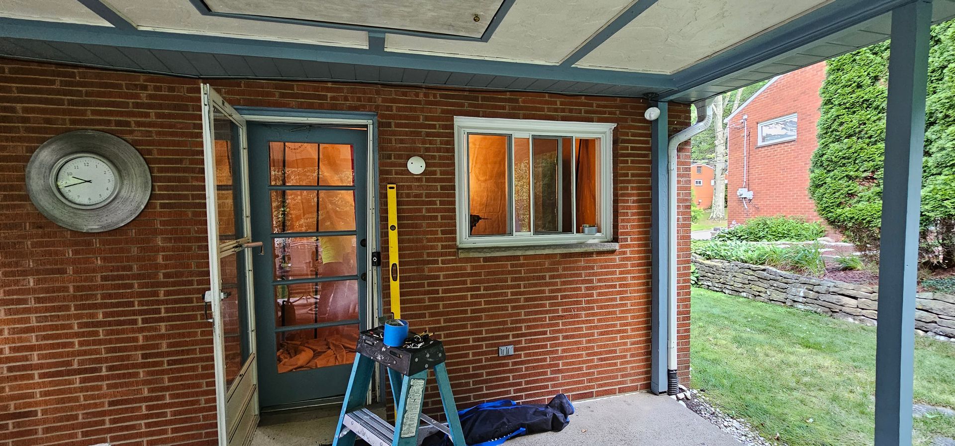 A brick-walled porch with a door, window, and clock on the wall. A green yard is visible on the right.