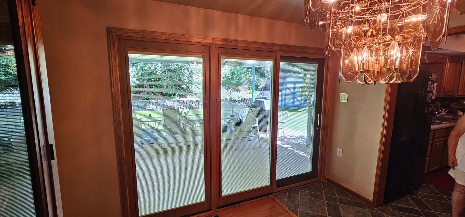 Three-panel sliding glass doors with a wooden frame lead to a backyard. A chandelier hangs above a tiled floor.