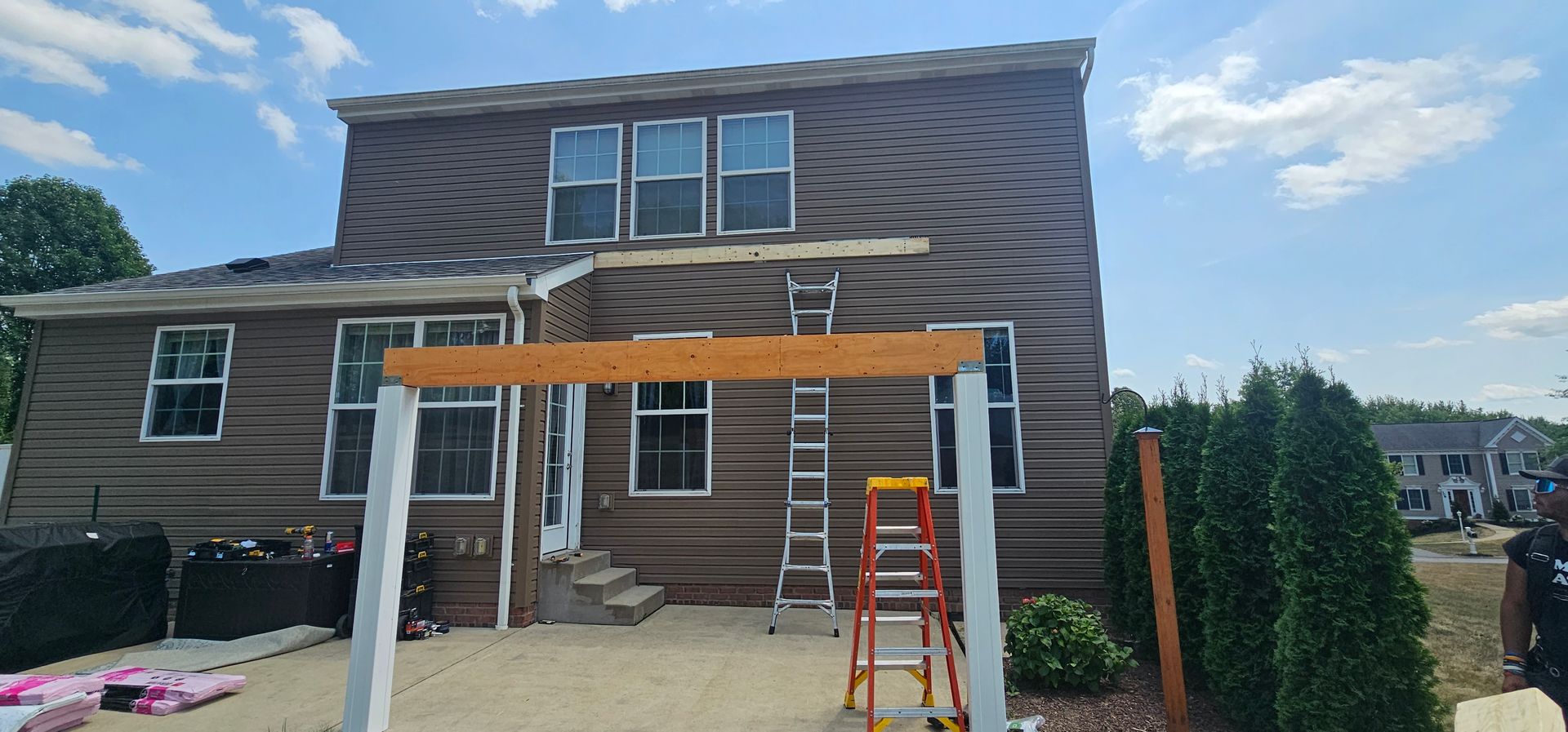 Back of a brown house under construction, with ladders and a partially built pergola. Sunny day.
