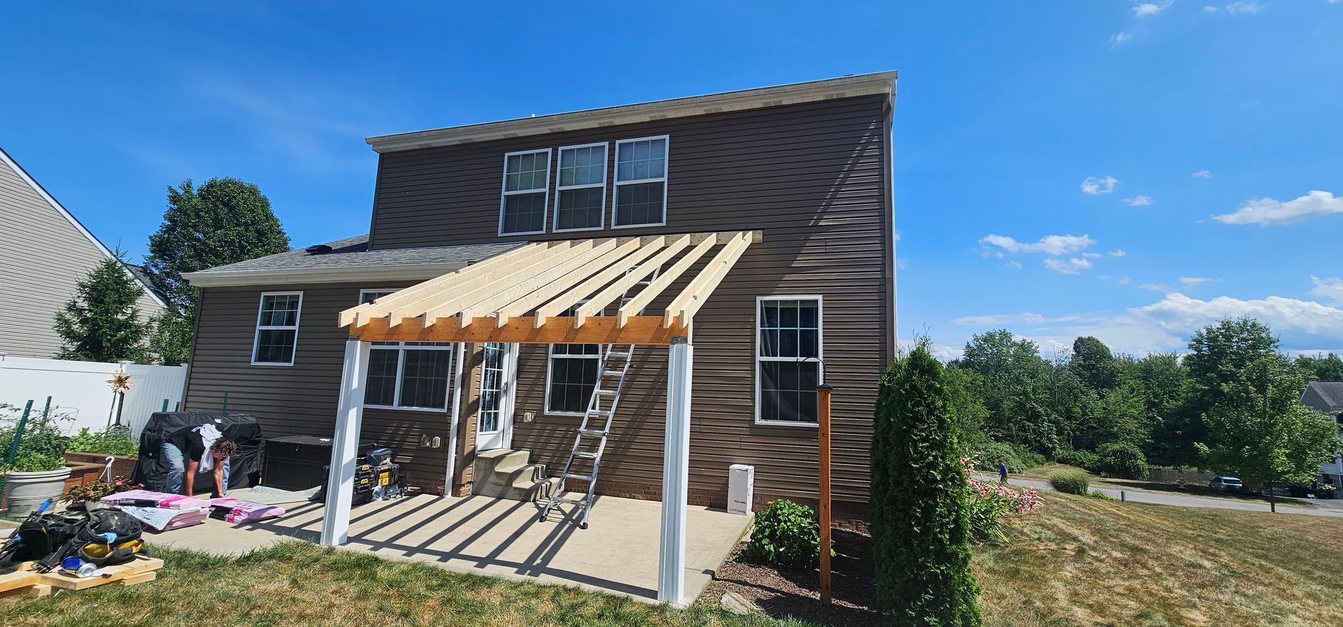 Back of a two-story brown house with a pergola under construction. Blue sky and green grass in the background.